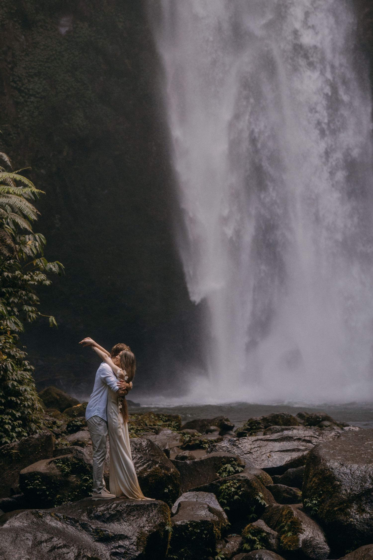 A person dances gracefully near a majestic waterfall surrounded by rocks and lush greenery in a scenic landscape.