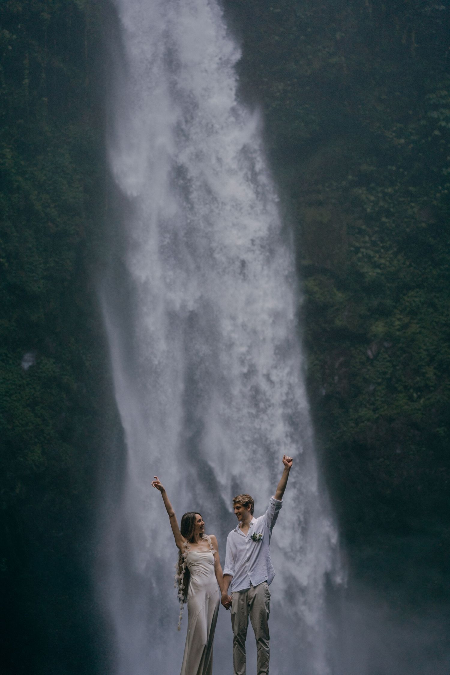 Two people in white outfits celebrate with raised arms in front of a majestic cascading waterfall.
