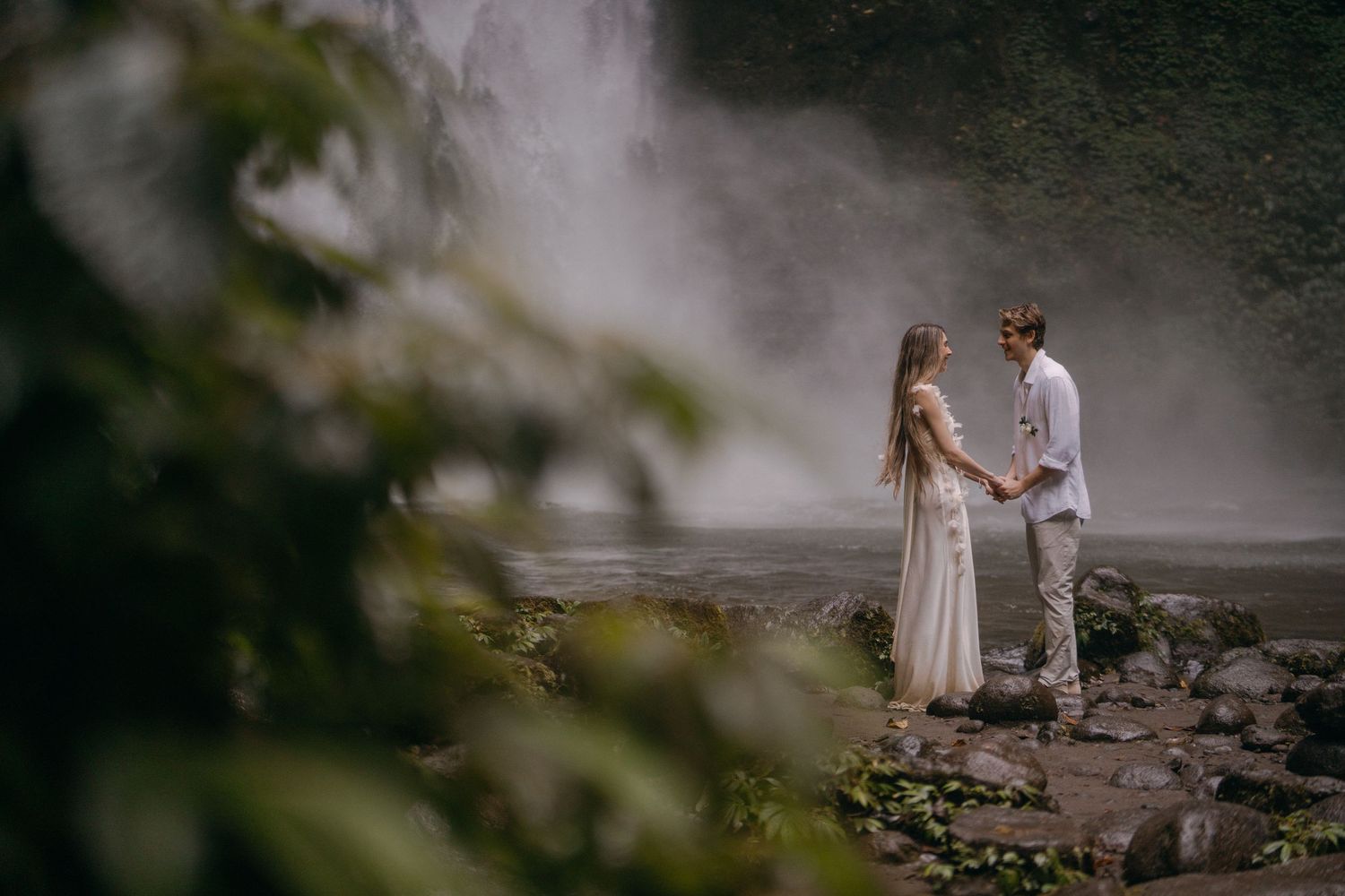 Two people in white outfits stand together near misty waterfall on rocky terrain during romantic photoshoot.