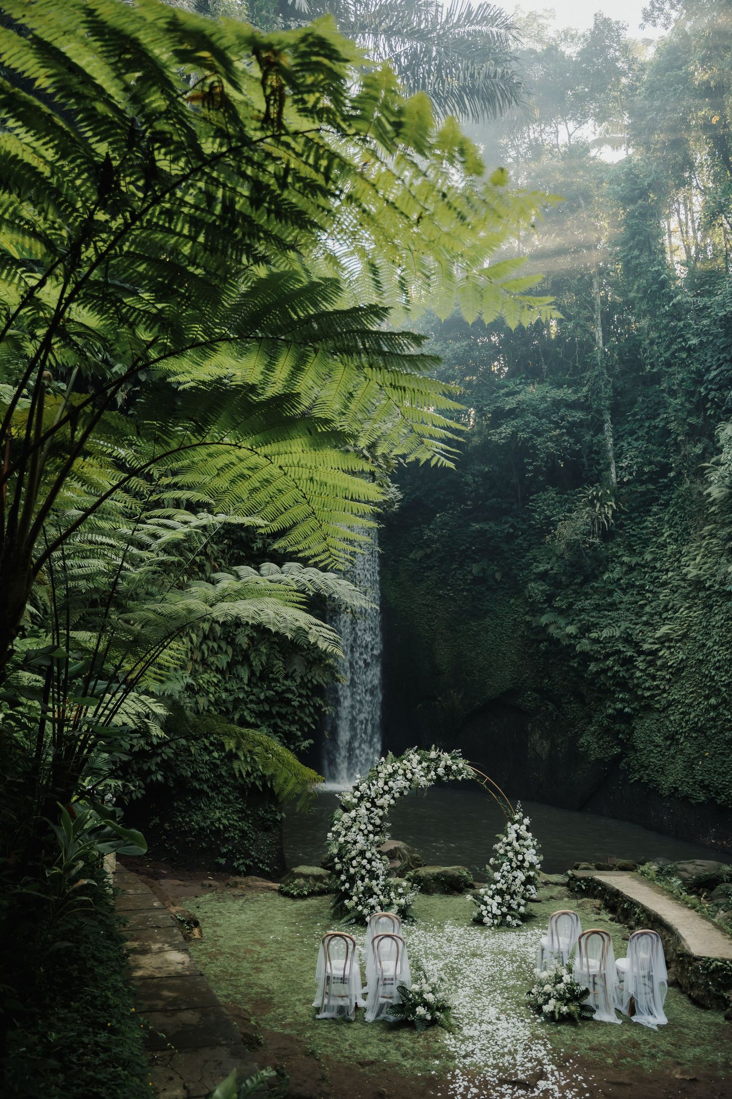 An elegant outdoor wedding ceremony setup with white floral arch before a tropical waterfall backdrop.