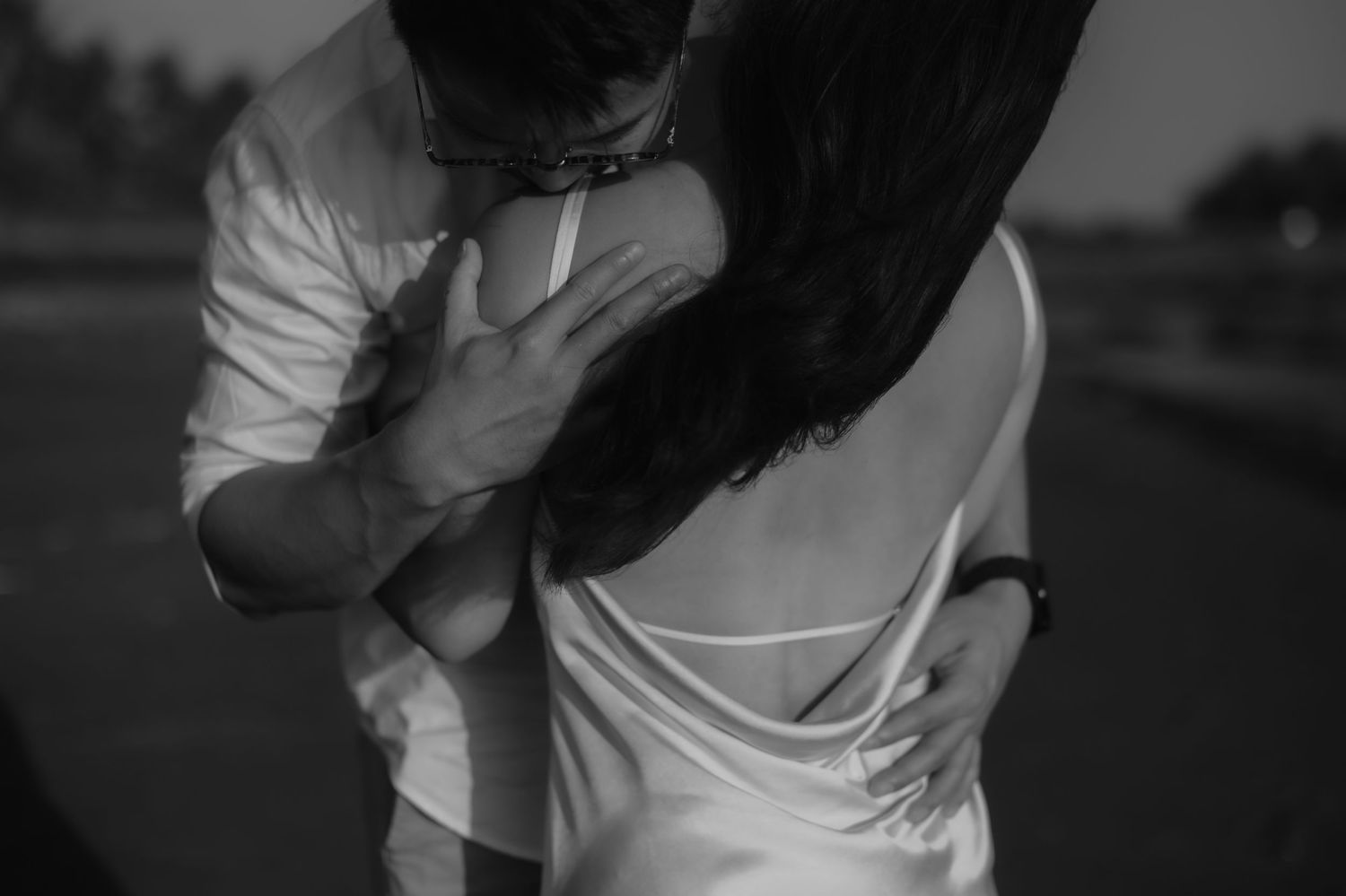 Black and white photo of a couple embracing romantically outdoors, man in white shirt hugging woman in backless dress.