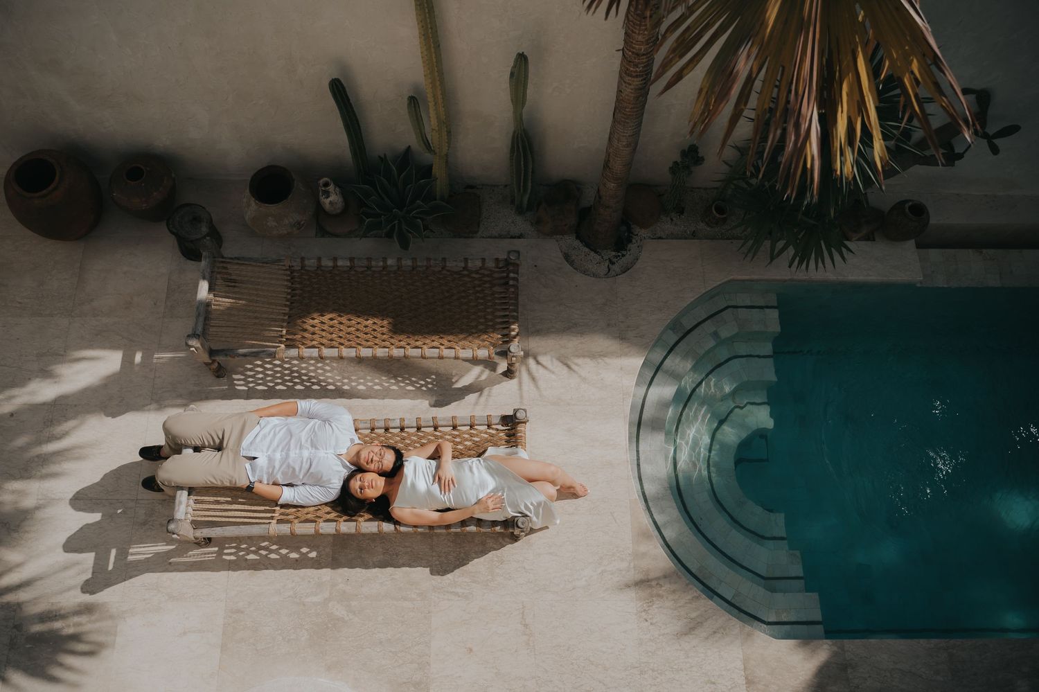 Aerial view of couple relaxing on wicker loungers beside a turquoise pool surrounded by tropical plants.