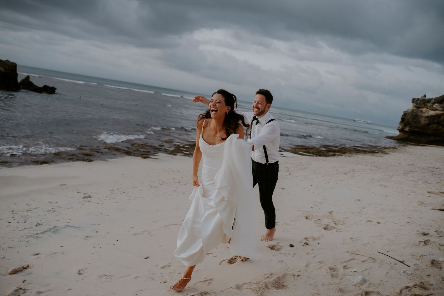 Wedding party members pose together on a sandy beach with moody gray skies in the background.