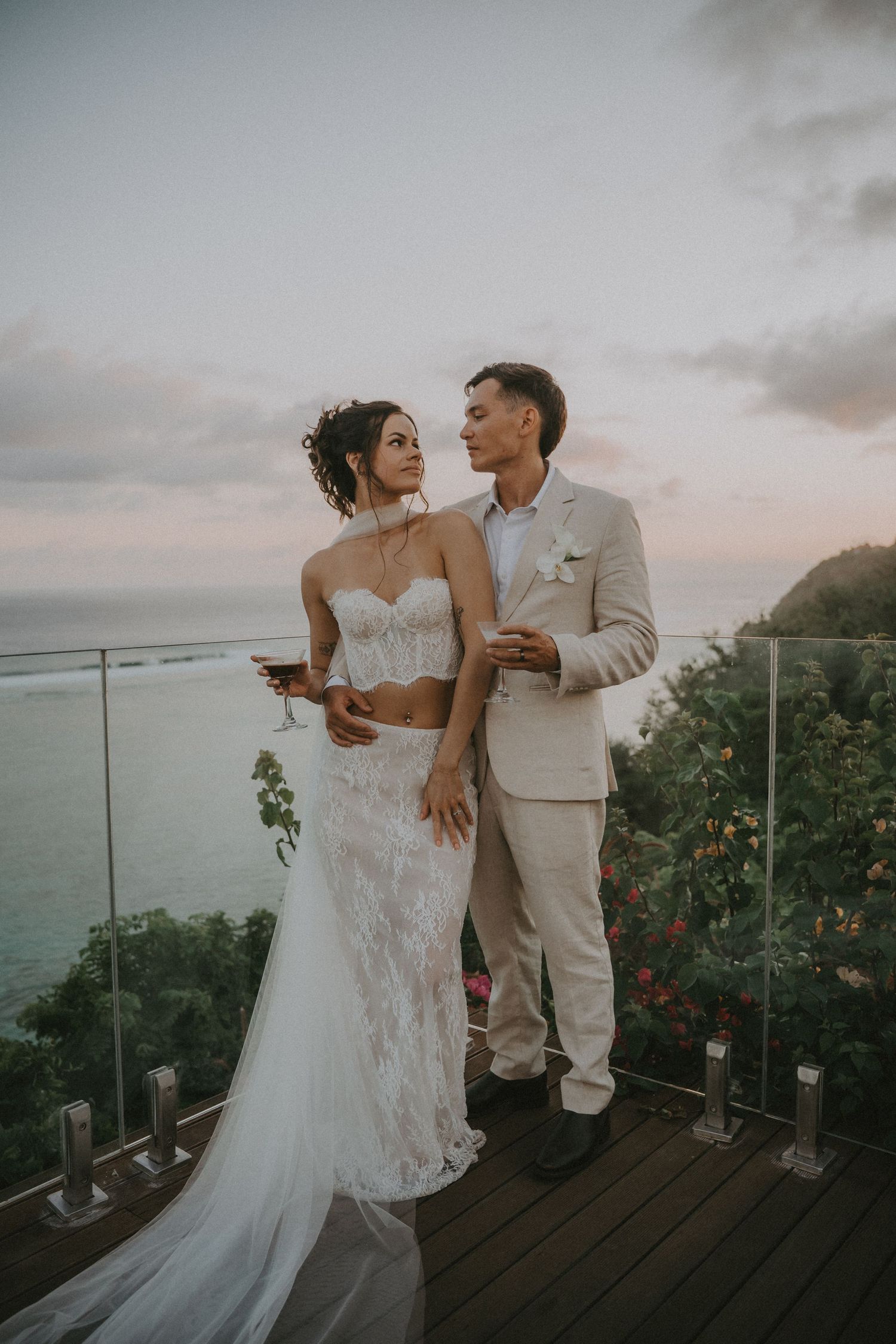 A romantic wedding couple embraces on an oceanfront balcony at sunset, with dramatic coastal views in the background.