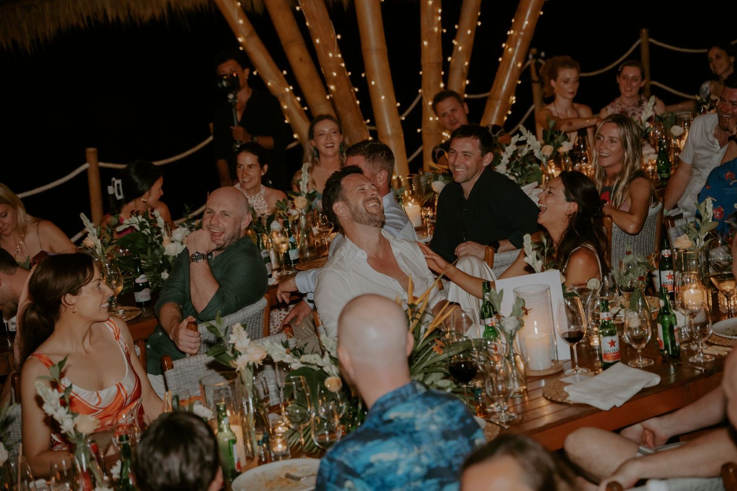 Wedding guests laugh and celebrate at a long reception table decorated with floral centerpieces and candles.