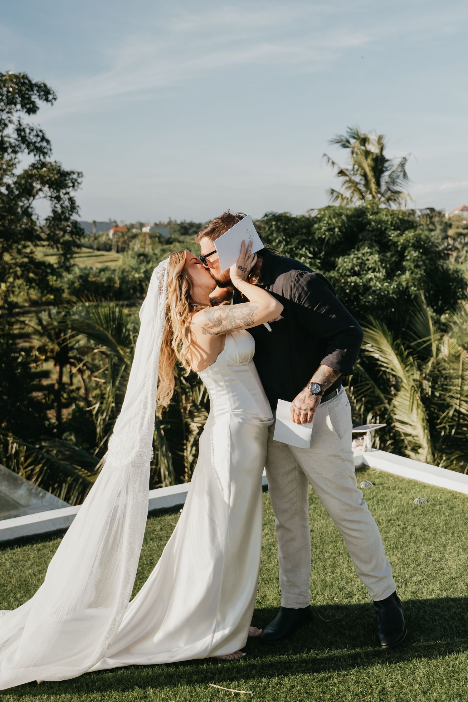 Bride and groom share first kiss at outdoor wedding ceremony with lush tropical greenery backdrop.