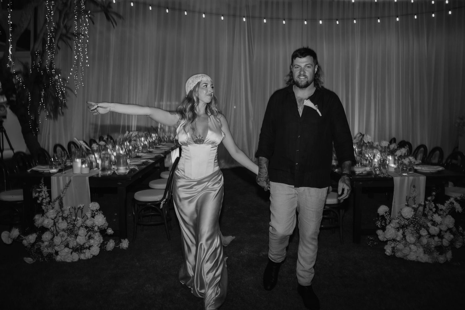 Bride and groom make grand entrance into elegantly lit reception hall with candles and floral centerpieces.