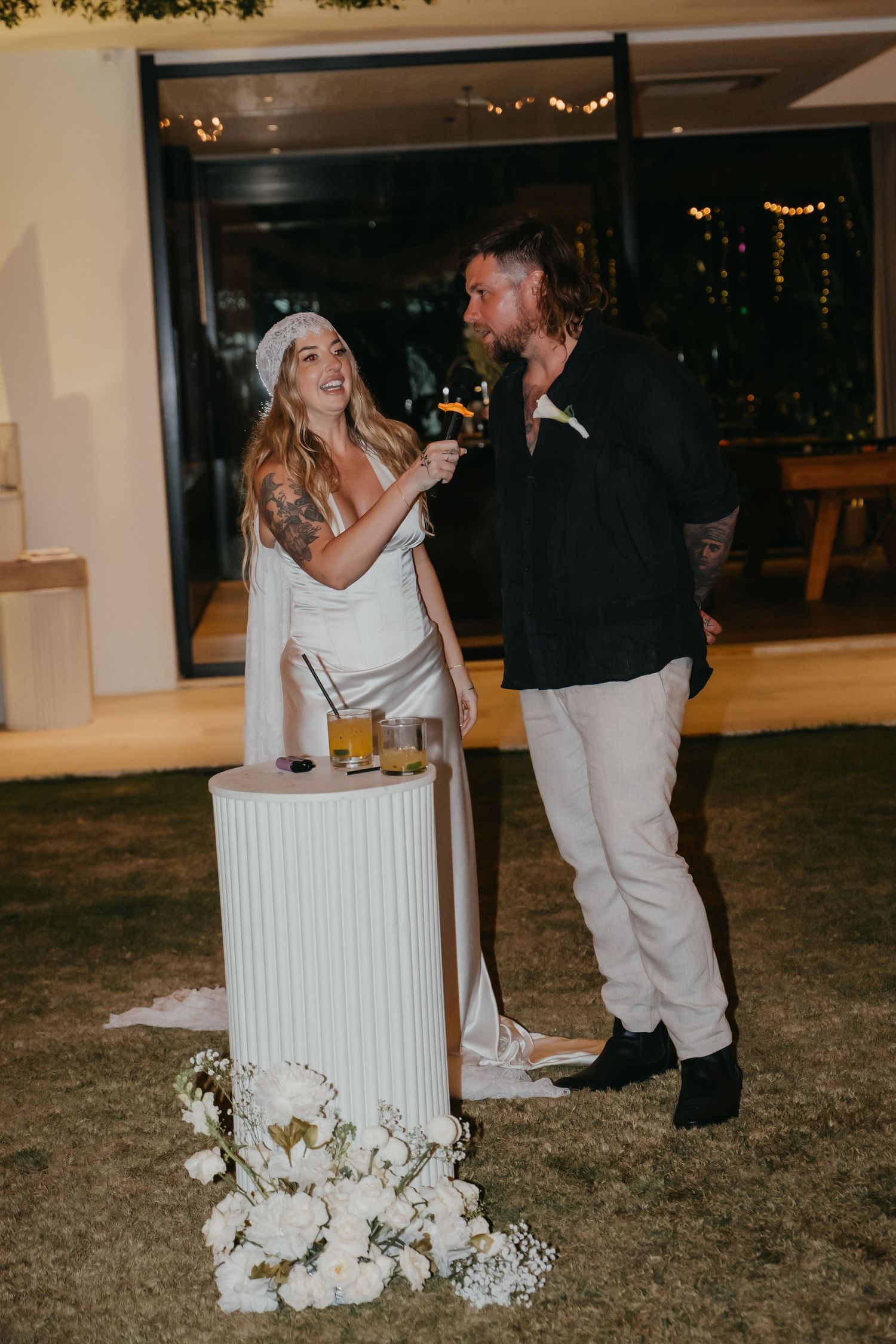 Bride and groom sharing a drink together at a white pedestal table decorated with white roses at their evening reception.