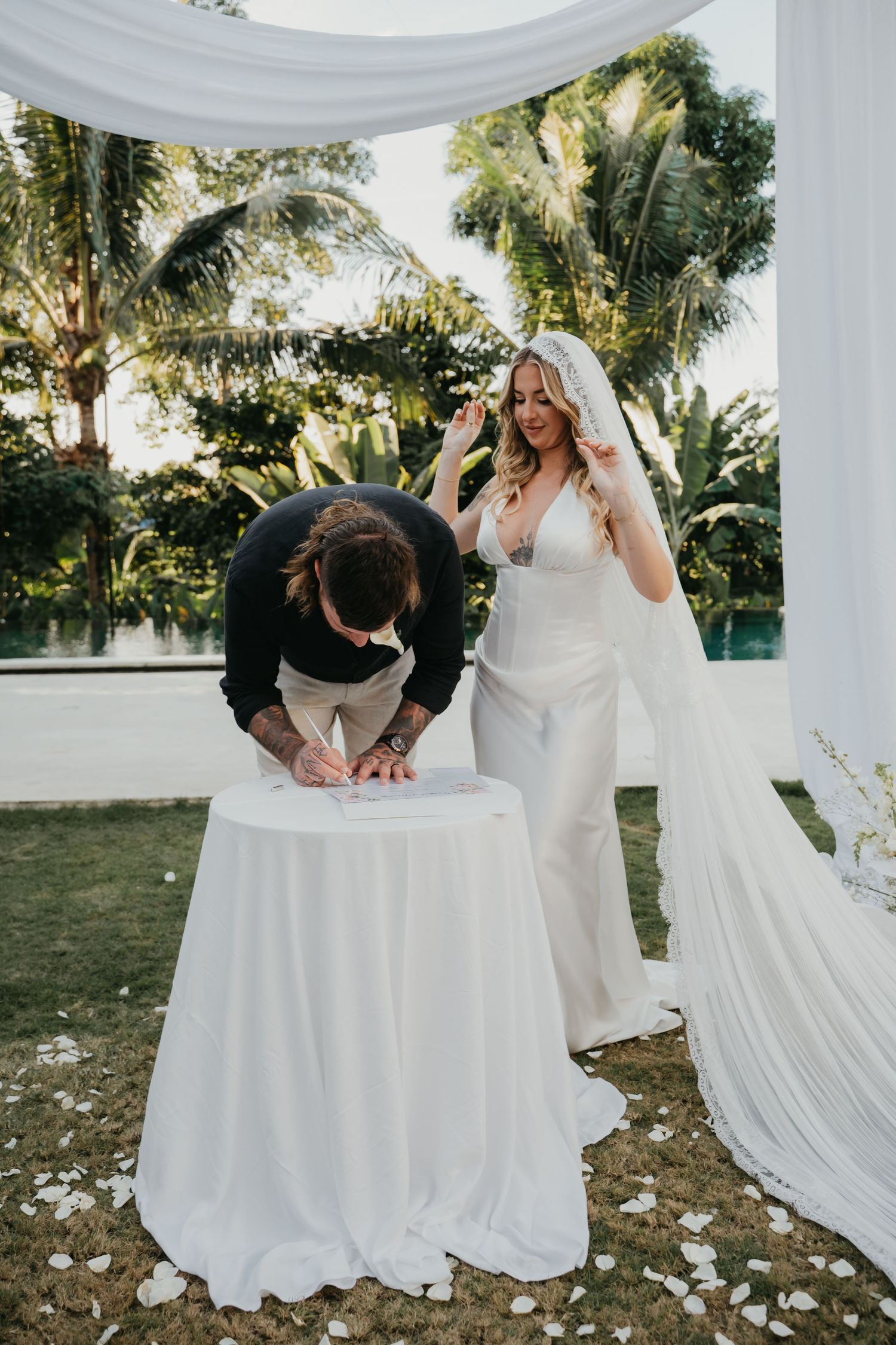 Groom signs wedding certificate at draped table while bride in white gown and veil raises her hand outdoors.