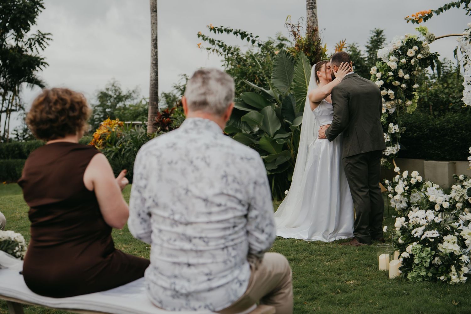 Couple sharing first kiss at white floral ceremony arch watched by seated wedding guests.