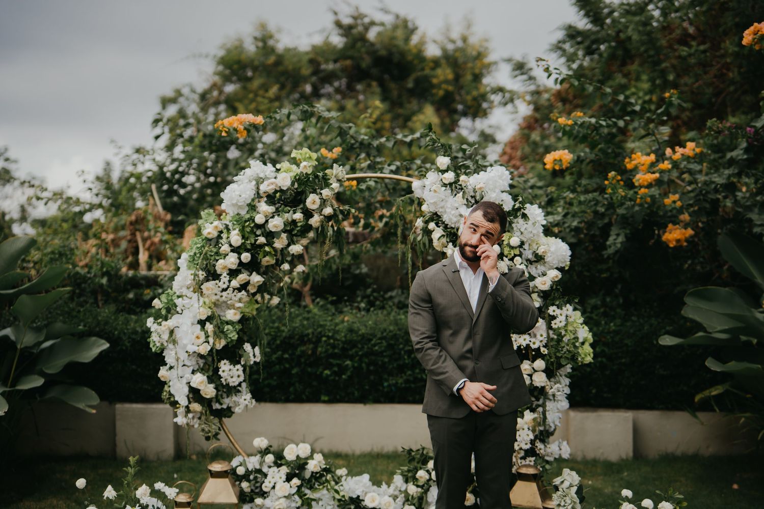Smiling groom in a grey suit standing before a circular floral arch with white flowers outdoors.