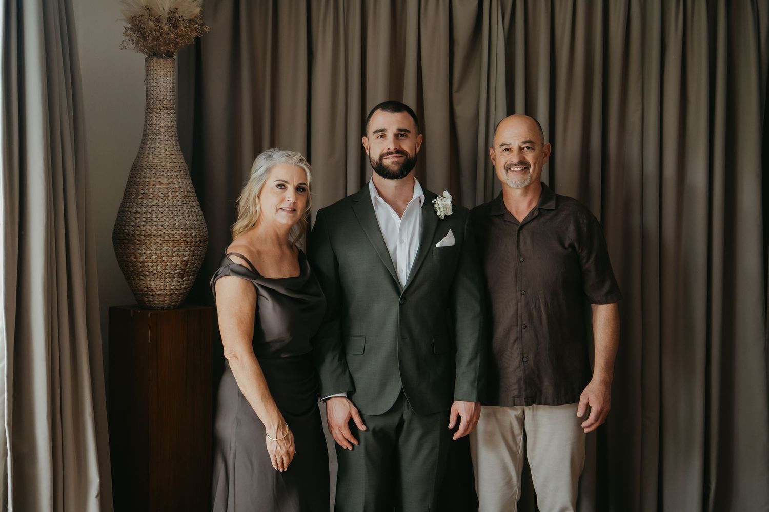 A groom in a dark suit poses with his parents in front of elegant gray curtains at a wedding.