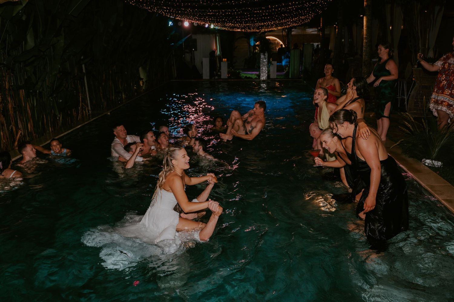 Bride and wedding guests enjoy a spontaneous late-night pool party under string lights at reception.