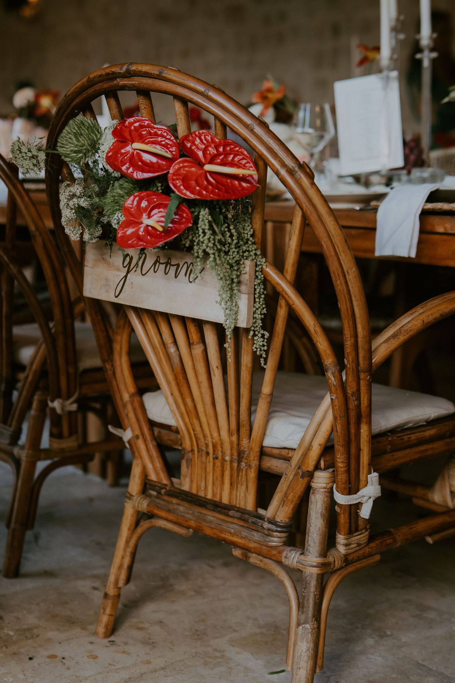 Wooden rattan groom chair decorated with red anthurium flowers and greenery at tropical wedding reception.