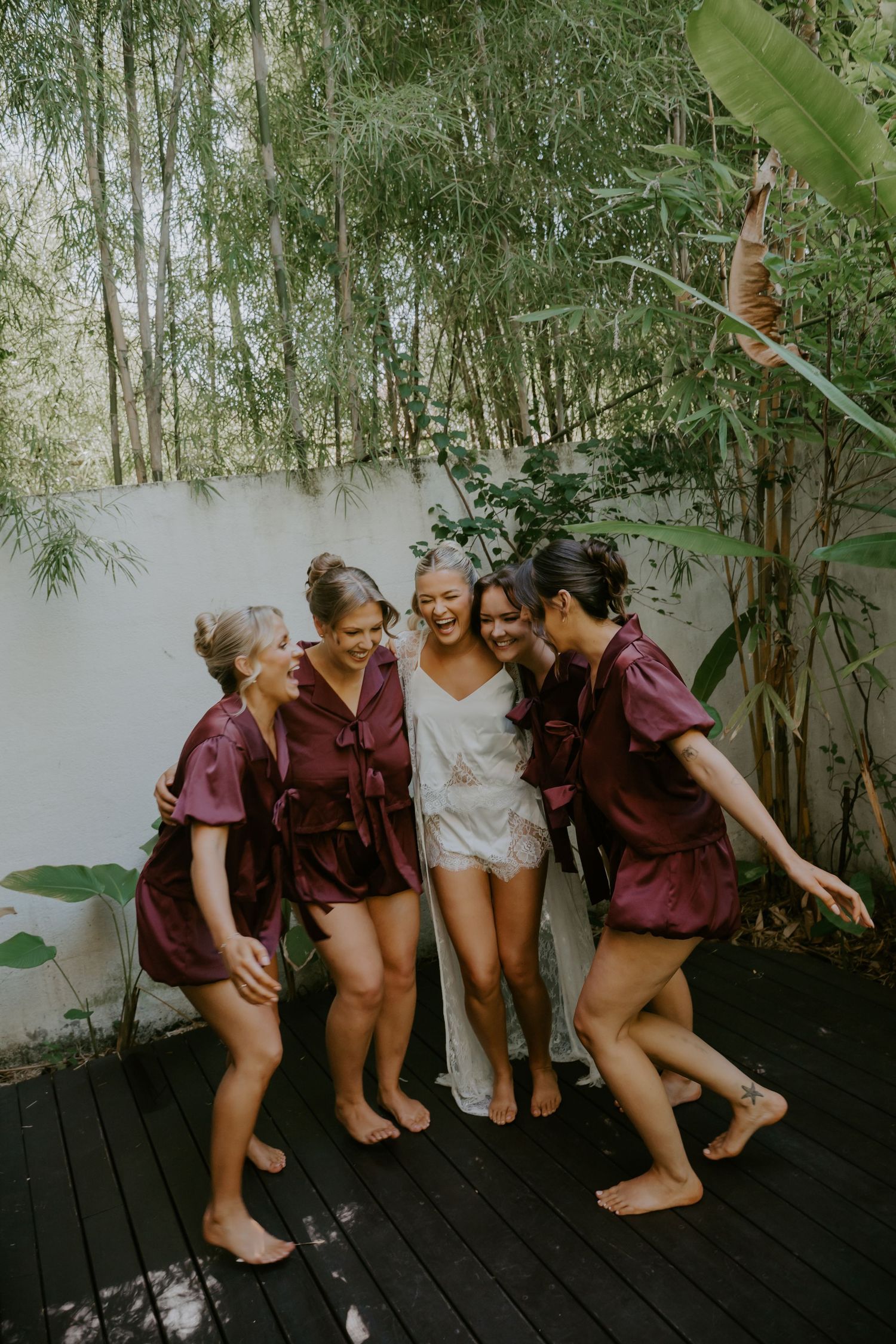 Bride in white slip surrounded by four bridesmaids in burgundy satin robes laughing together on tropical outdoor deck.