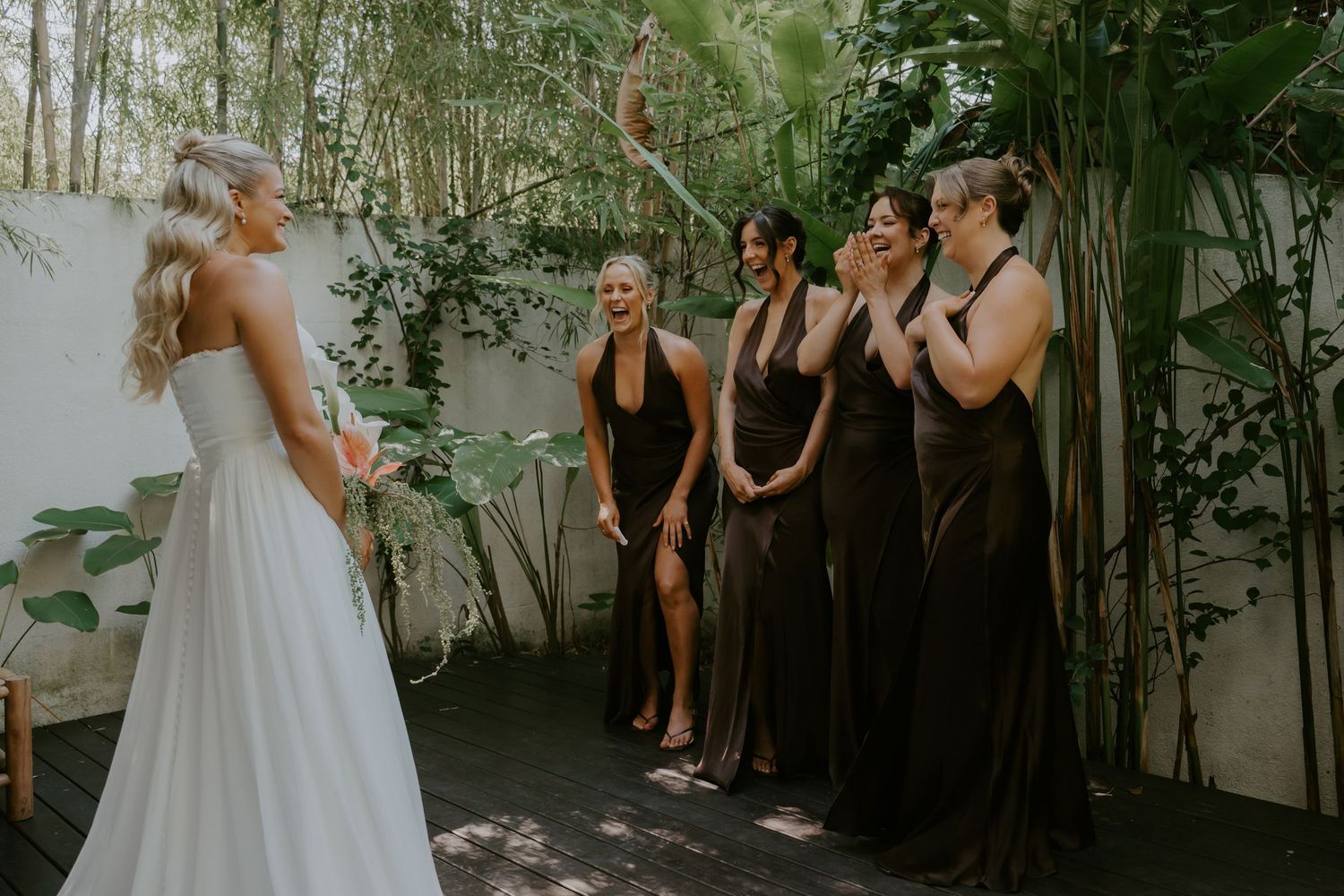Bridesmaids in dark brown halter gowns react with joy as bride reveals her wedding dress outdoors.