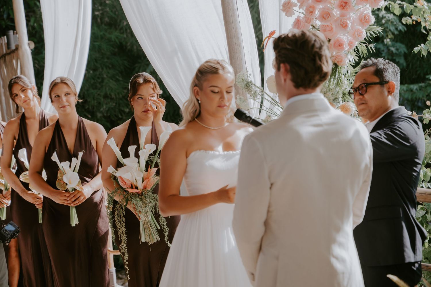 Bride in strapless white gown reads vows while bridesmaids in brown dresses stand beside her.
