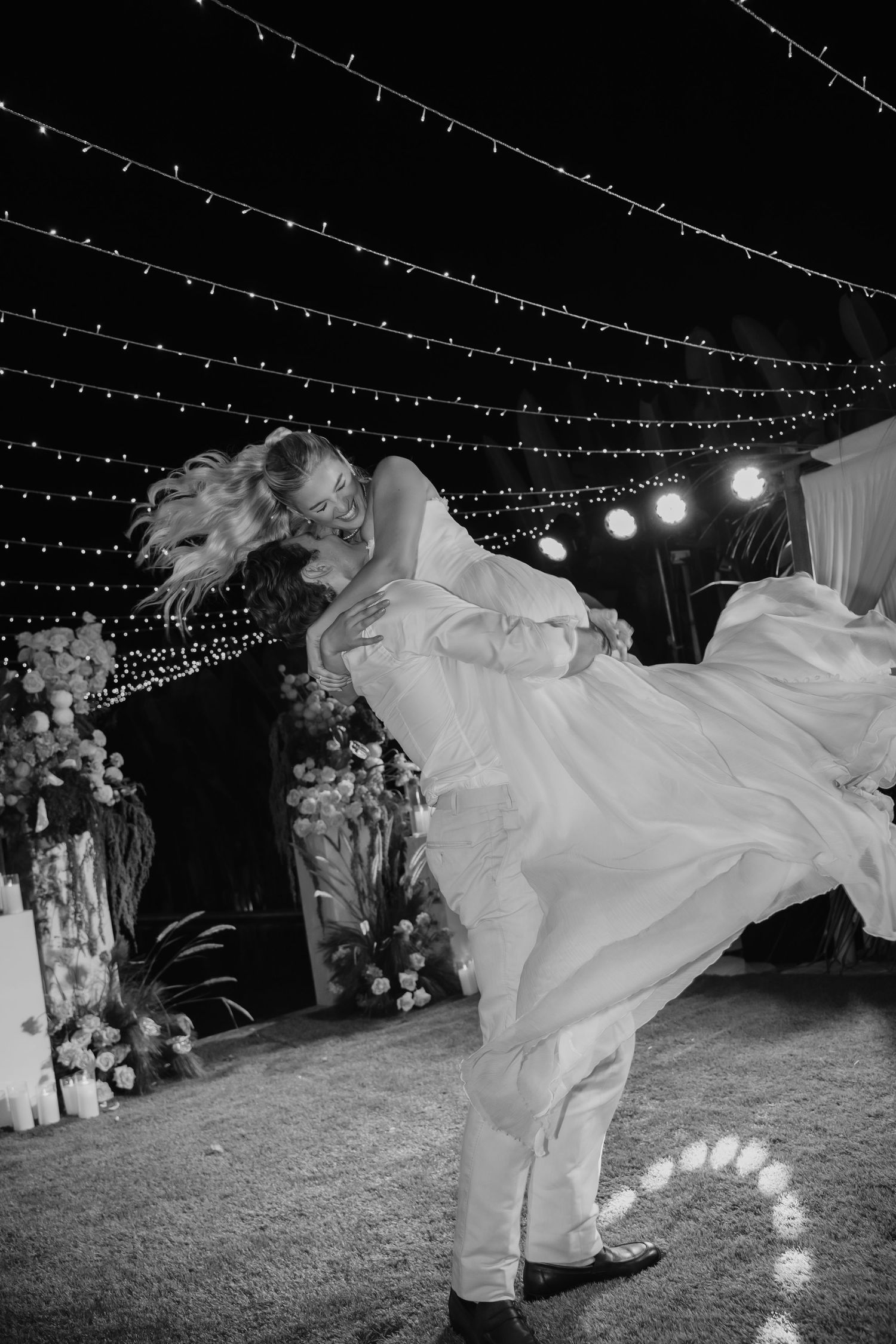 Black and white photo of groom lifting bride in flowing gown during energetic first dance at night.