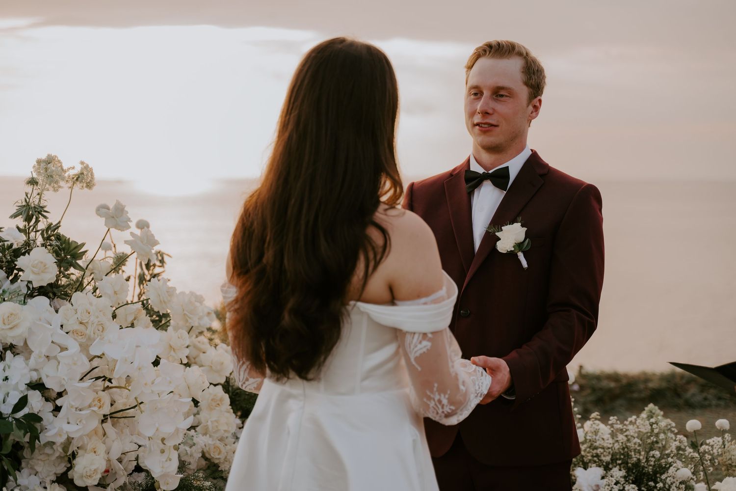 Groom in burgundy tuxedo holds brides hands at romantic sunset wedding ceremony with white floral arrangements.