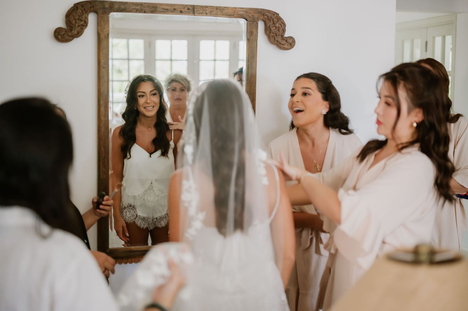 Bride in wedding dress and veil admired by bridesmaids in white robes reflected in ornate mirror.