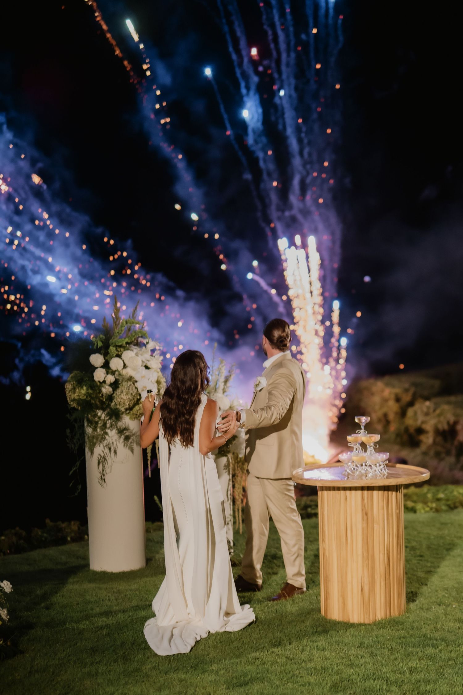 Bride and groom watch colorful fireworks display together beside champagne tower at outdoor wedding.