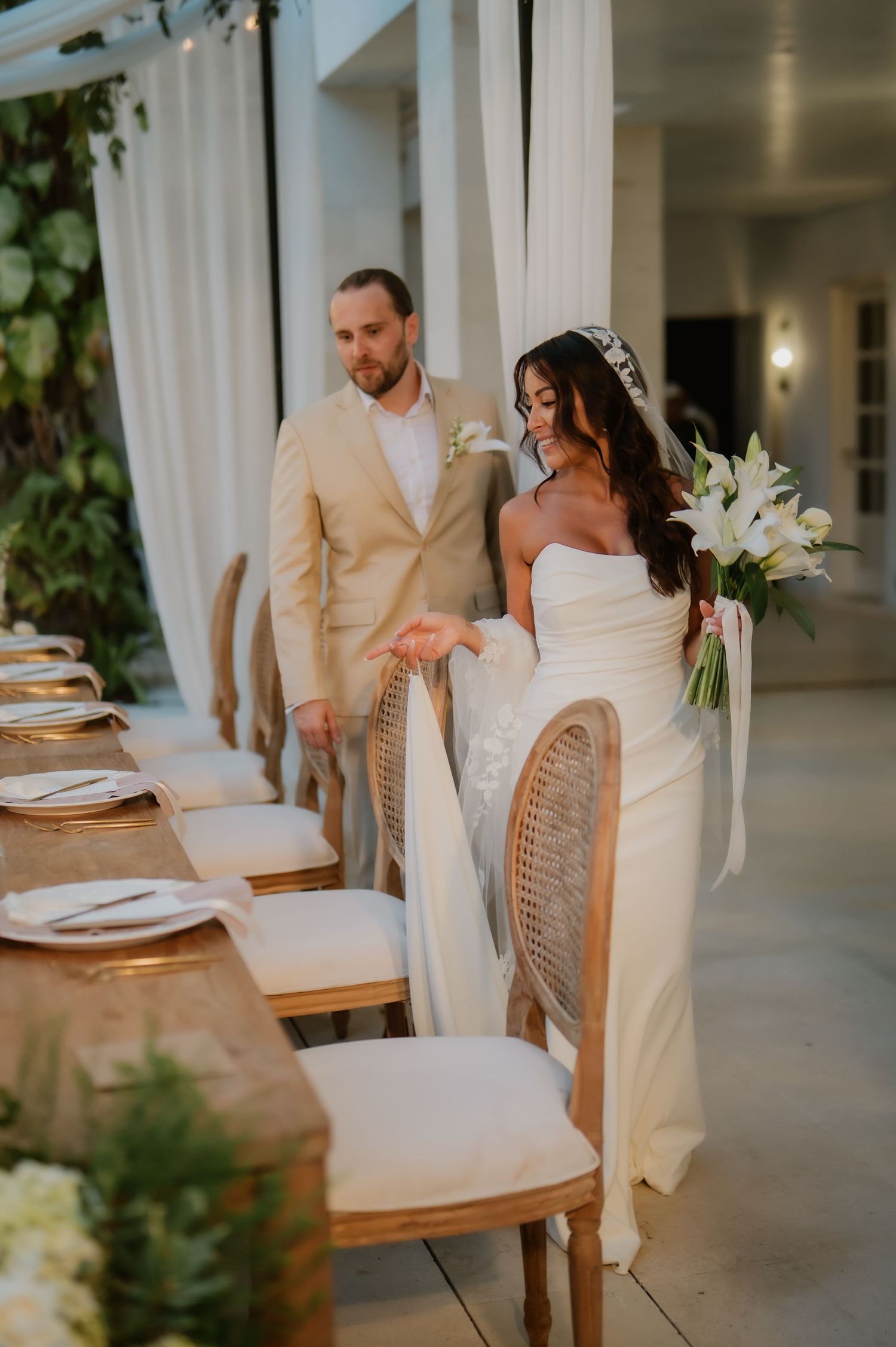 Bride in strapless gown holds white lily bouquet as groom leads her past elegant reception table.