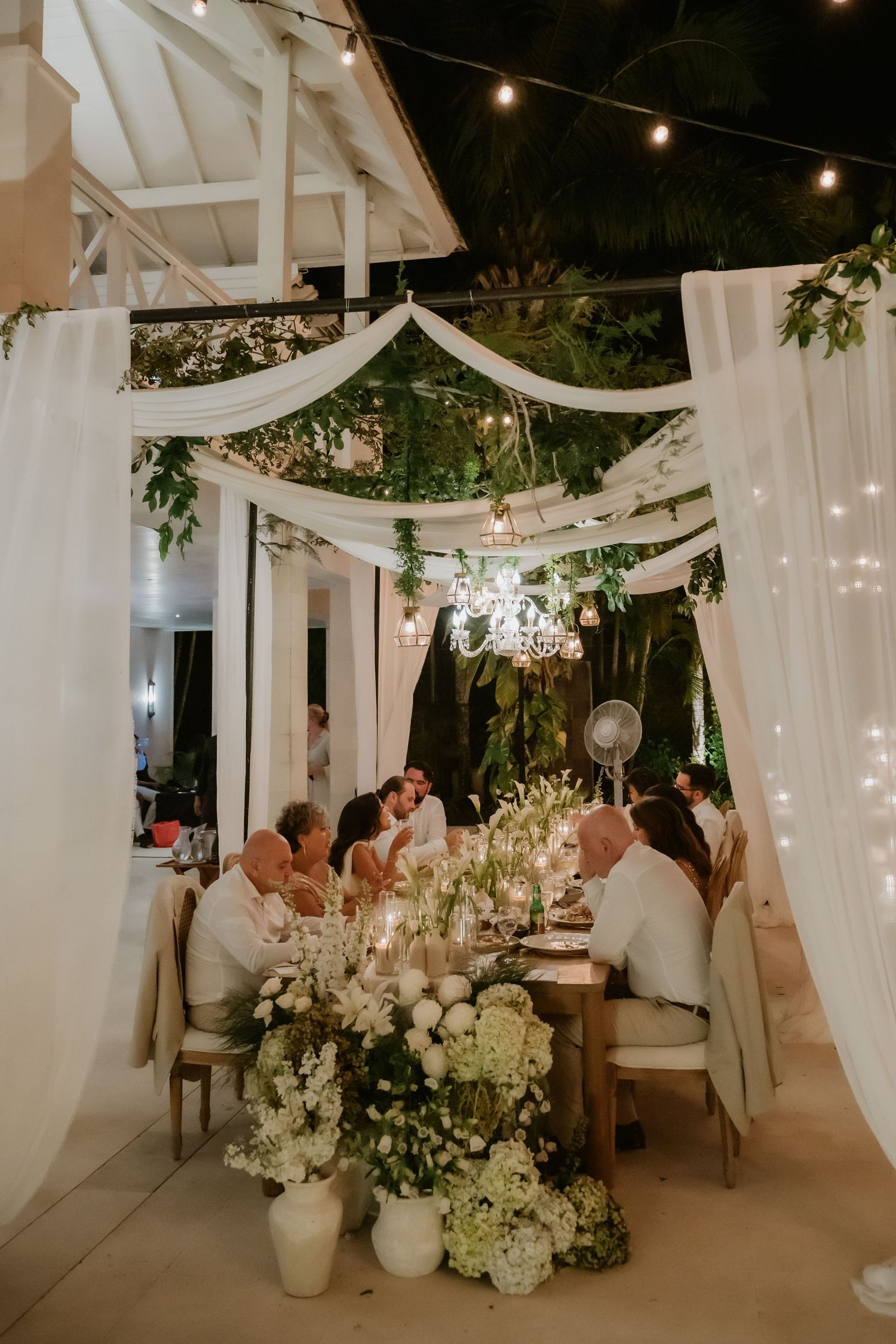 Guests dine at candlelit wedding reception table with white floral arrangements under draped fabric ceiling.