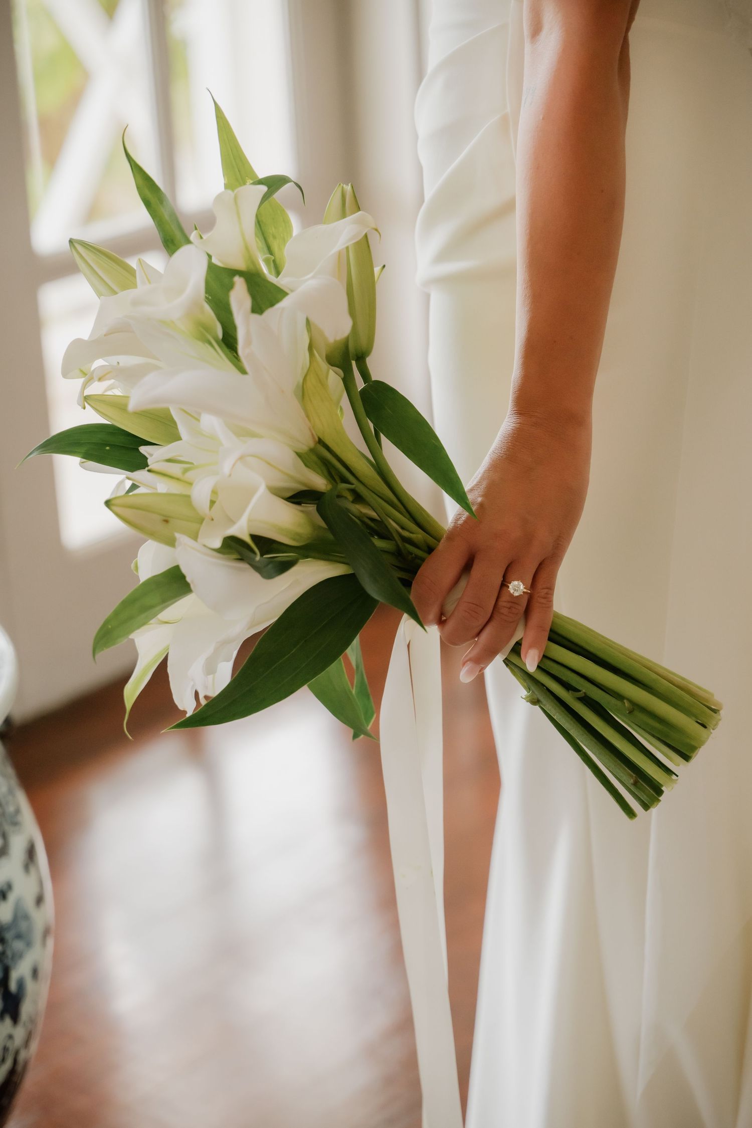 Bride holding a white lily wedding bouquet with green leaves while wearing an engagement ring and white gown.