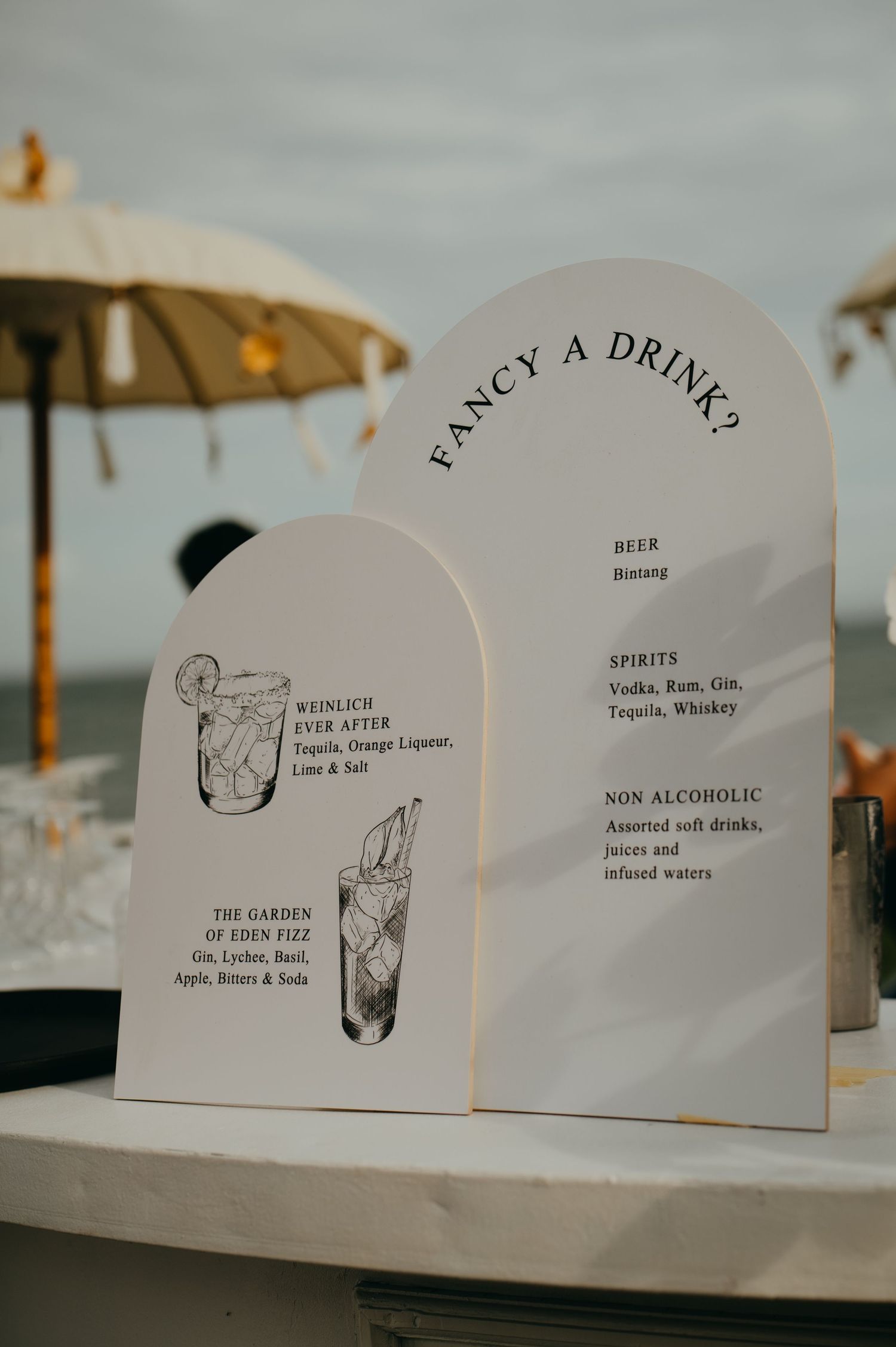 Elegant arched wedding drink menu signs reading Fancy A Drink displayed at an outdoor beach bar.