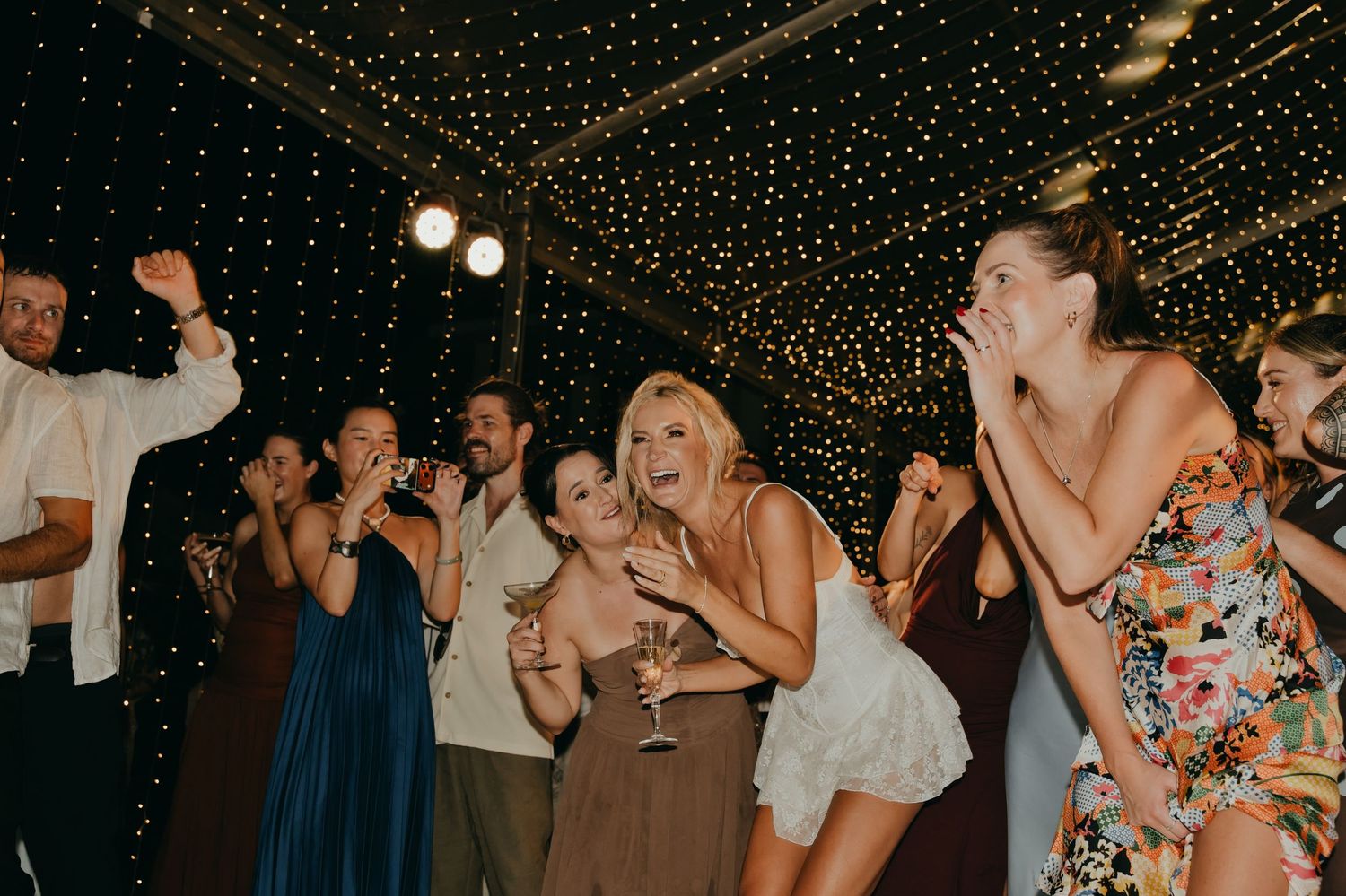 Bride and wedding guests laugh and dance together under twinkling lights at the reception party.