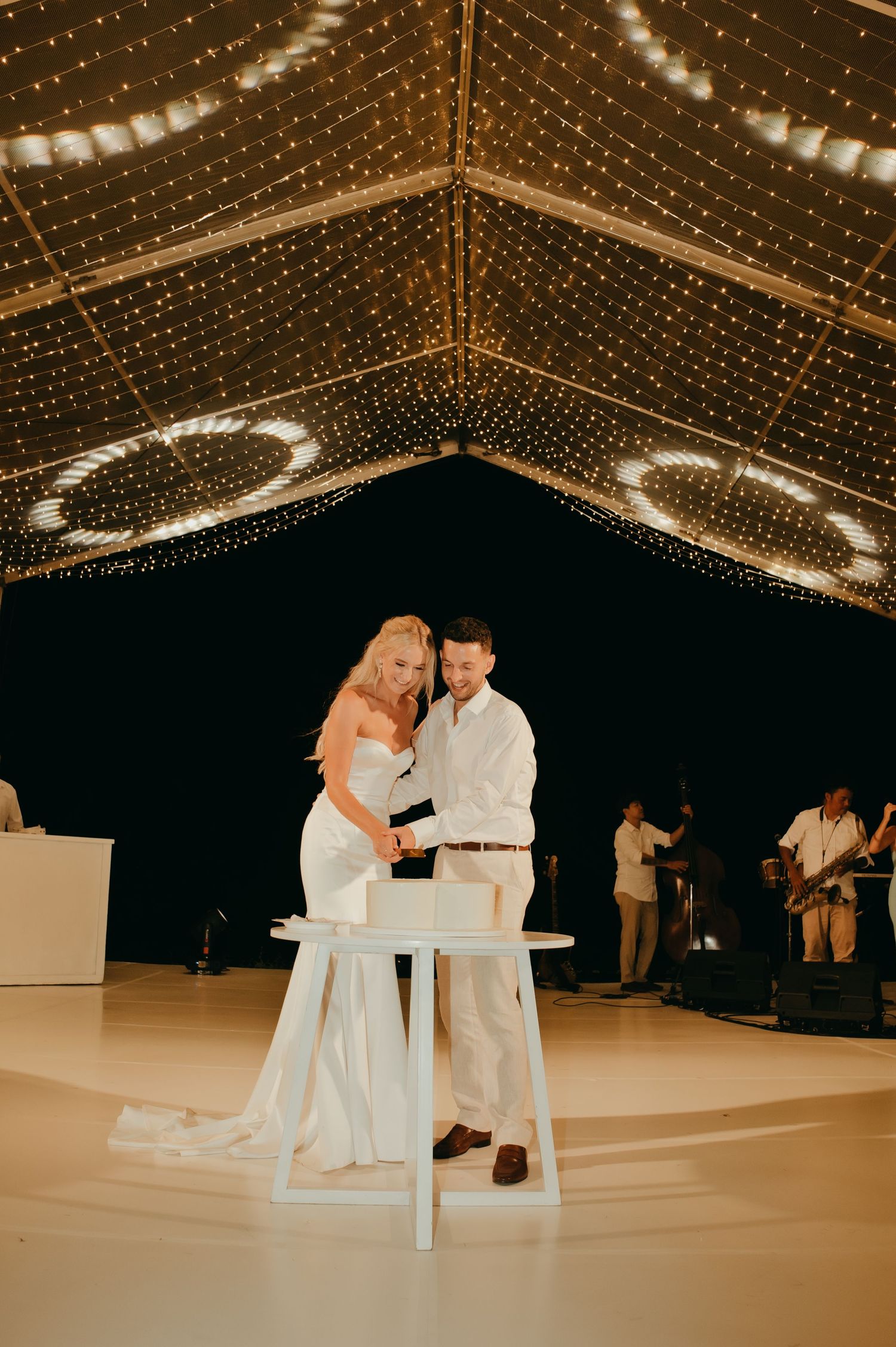 Bride and groom cutting white wedding cake under glowing fairy light canopy at night.