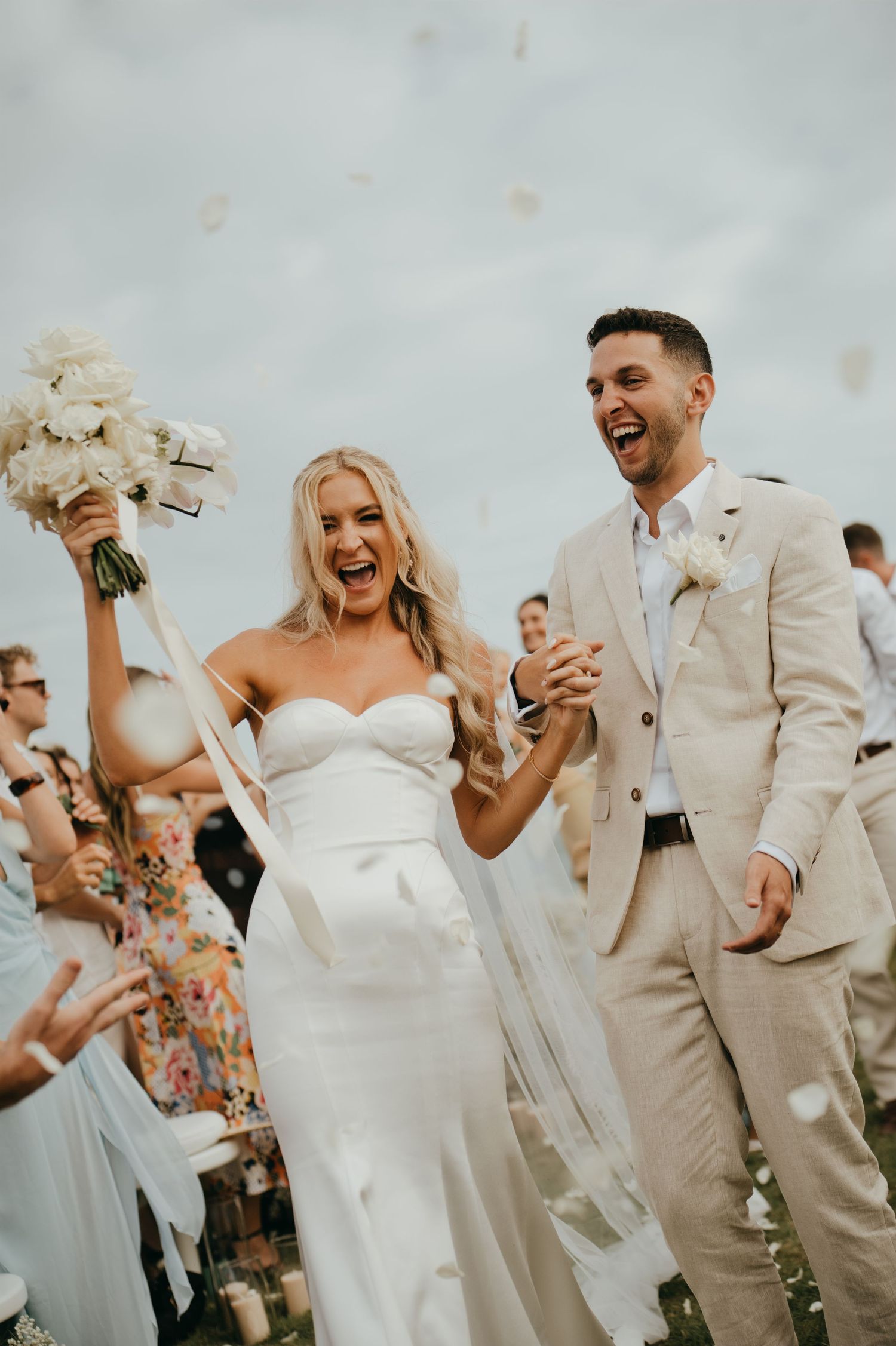 Joyful newlyweds walk down aisle as guests toss white flower petals at outdoor wedding.