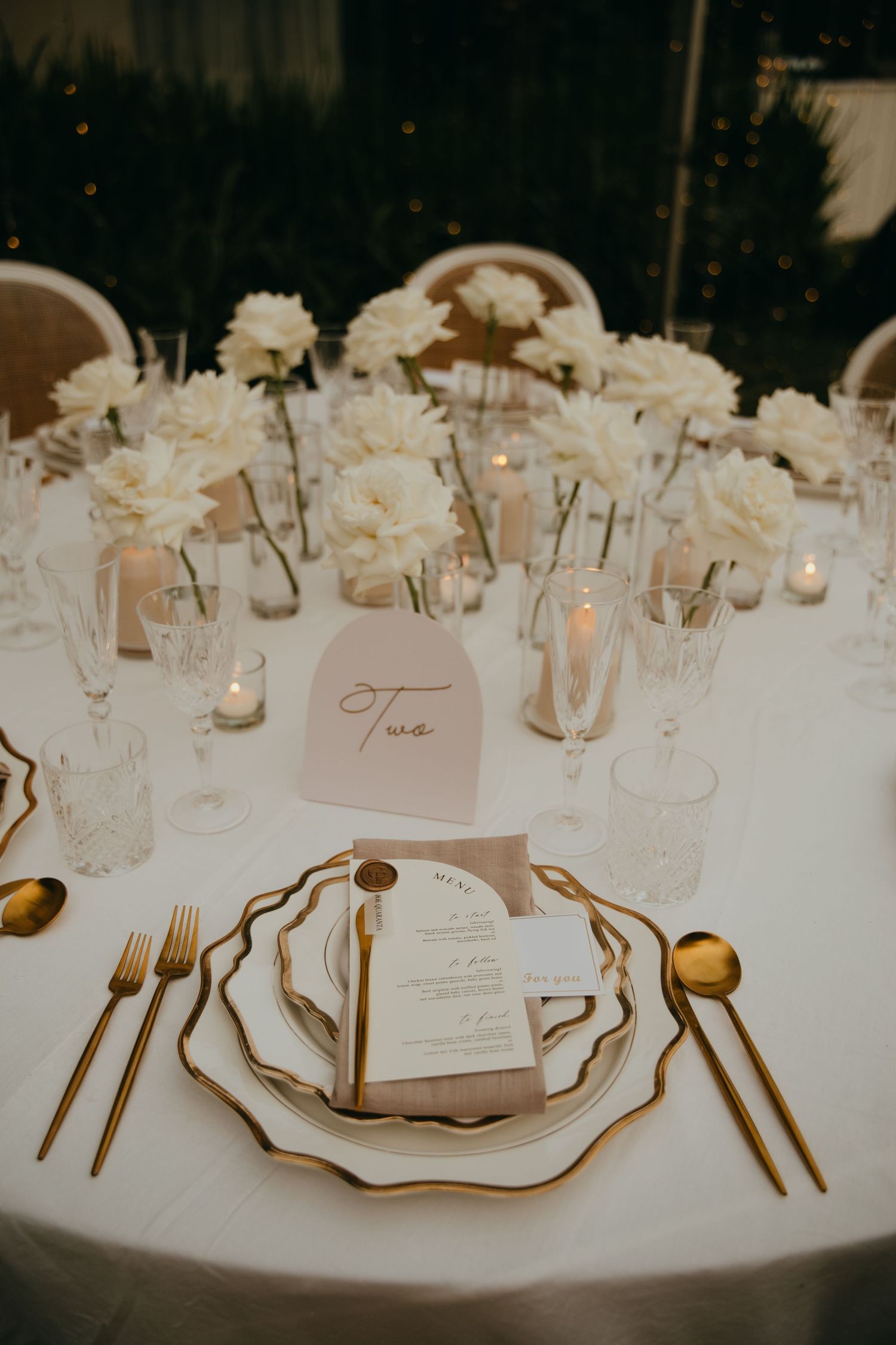 Elegant wedding table setting with gold cutlery, white florals, candles, and a table number sign.