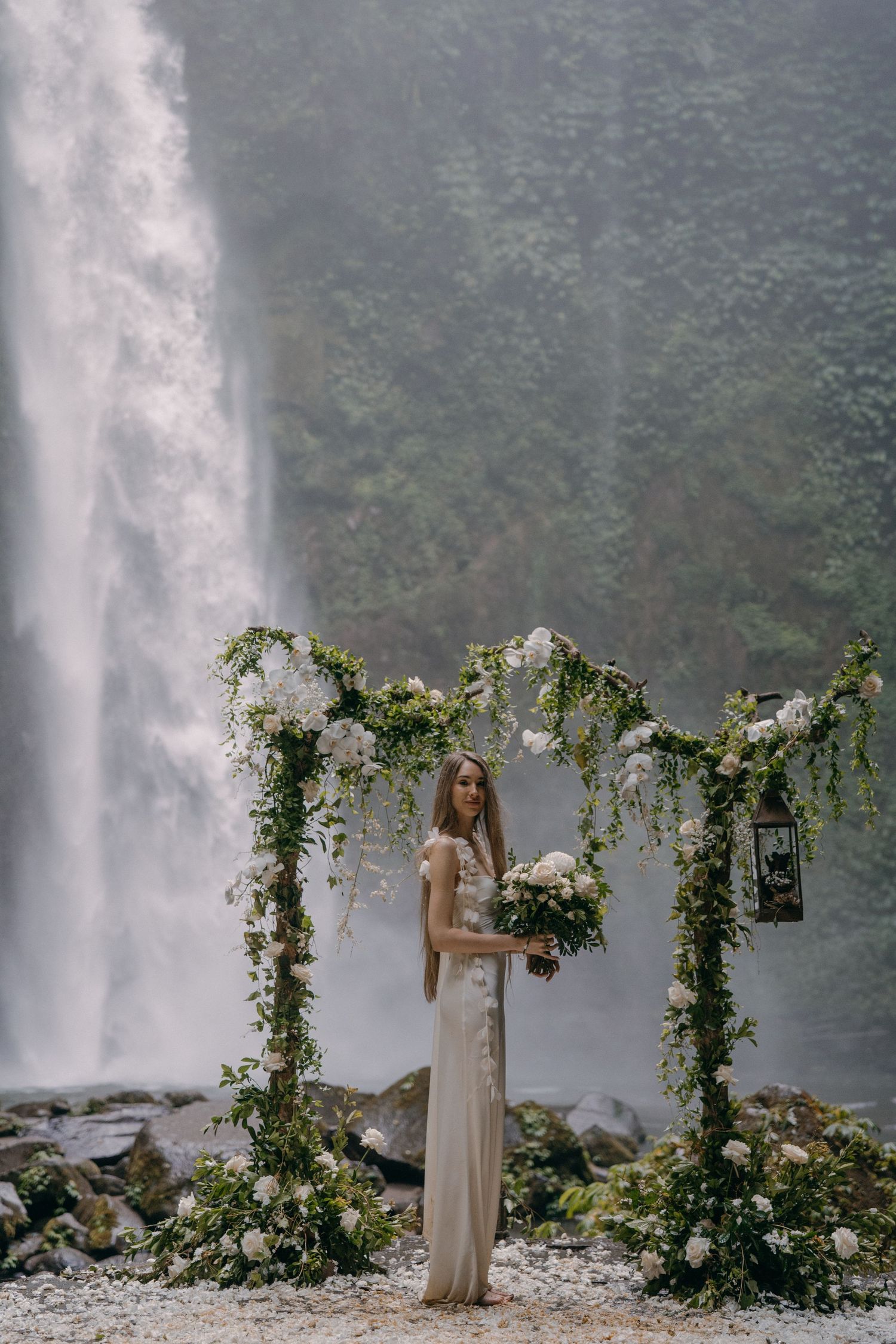 A person in a flowing dress poses near a majestic waterfall surrounded by floral archways in a dreamy forest setting.