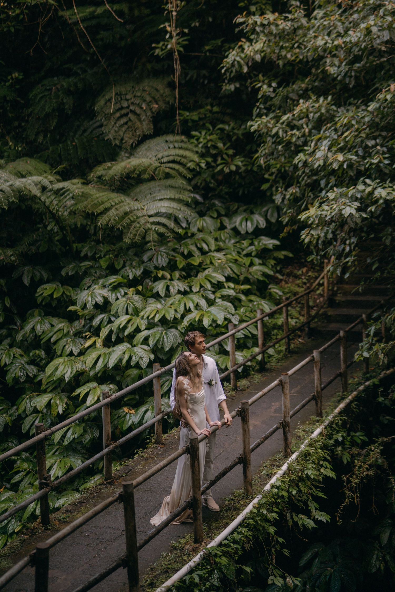 A couple walks along a wooden bridge pathway surrounded by lush tropical foliage in a romantic forest setting.