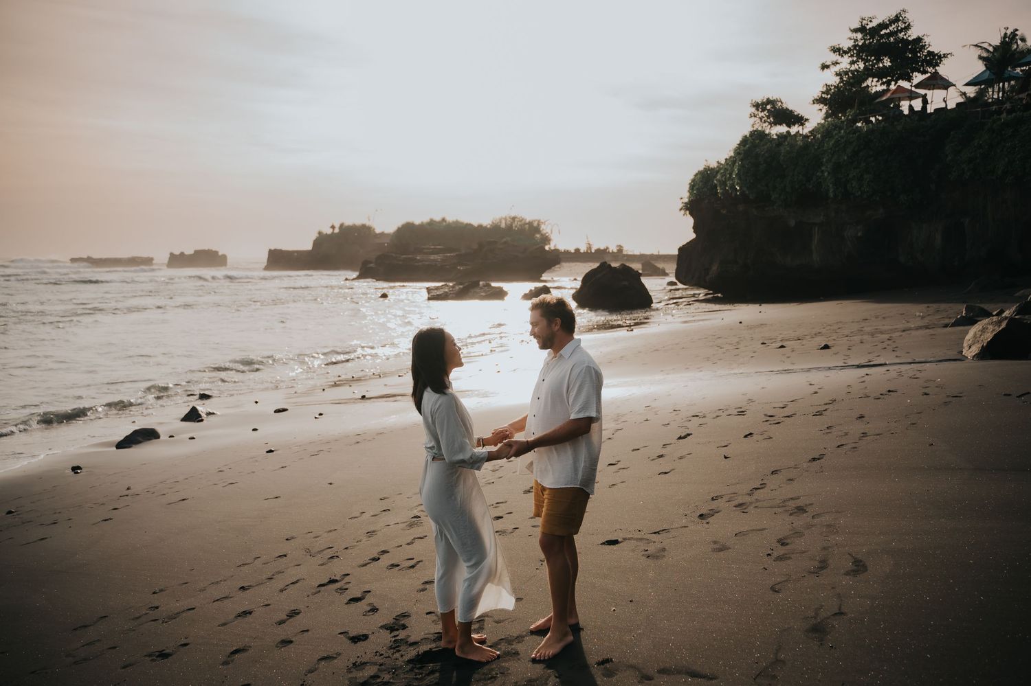 Couple walking along a misty beach at sunset with their footprints trailing behind in the sand.