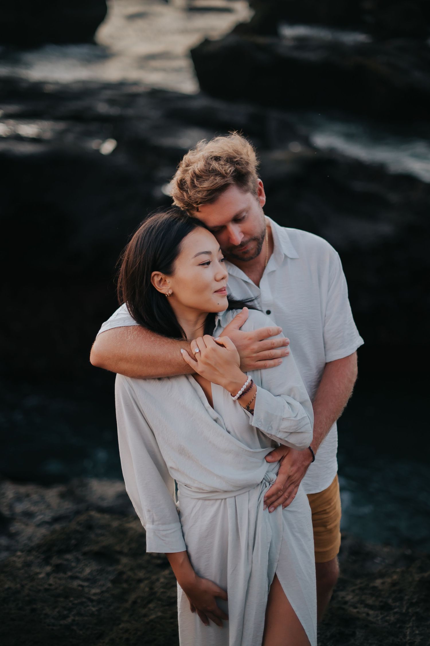 A romantic couple embraces on dark rocky cliffs during sunset, with the woman wearing a flowing white dress.