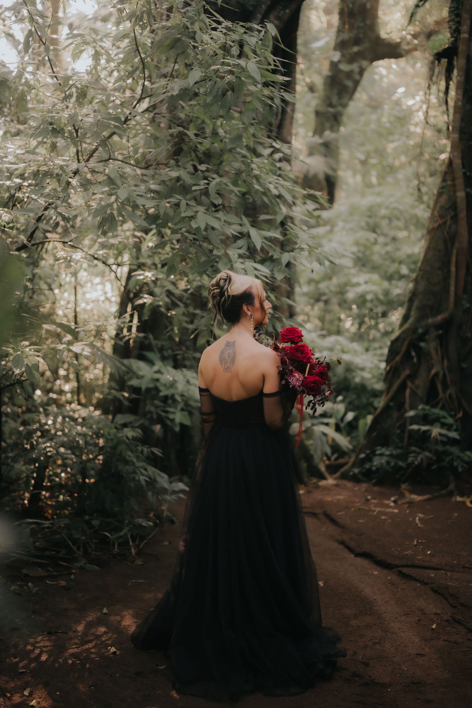 Back view of woman in black dress holding red roses while standing on forest path.