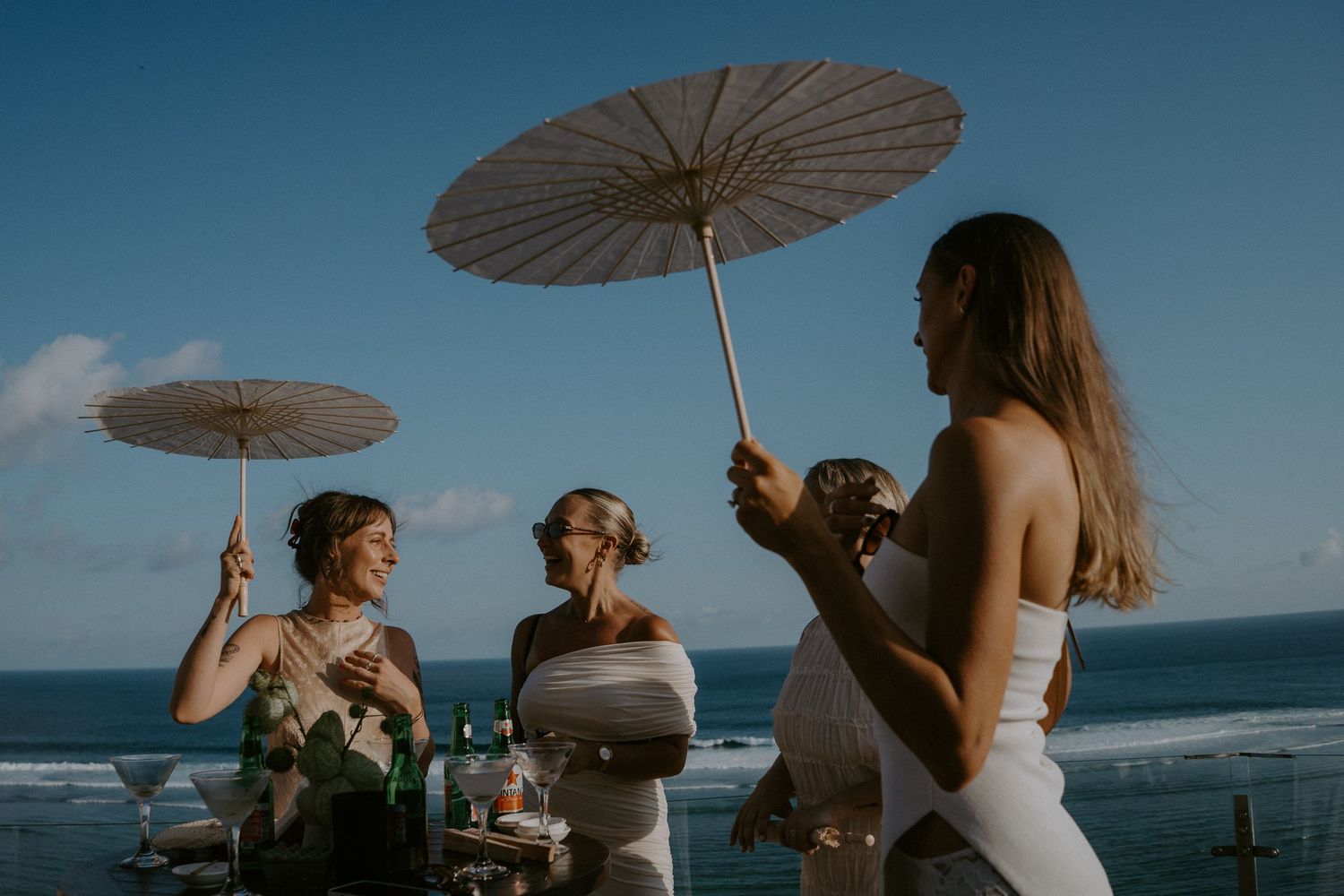 Wedding party holds white parasols while posing against blue ocean backdrop at sunset beach ceremony.