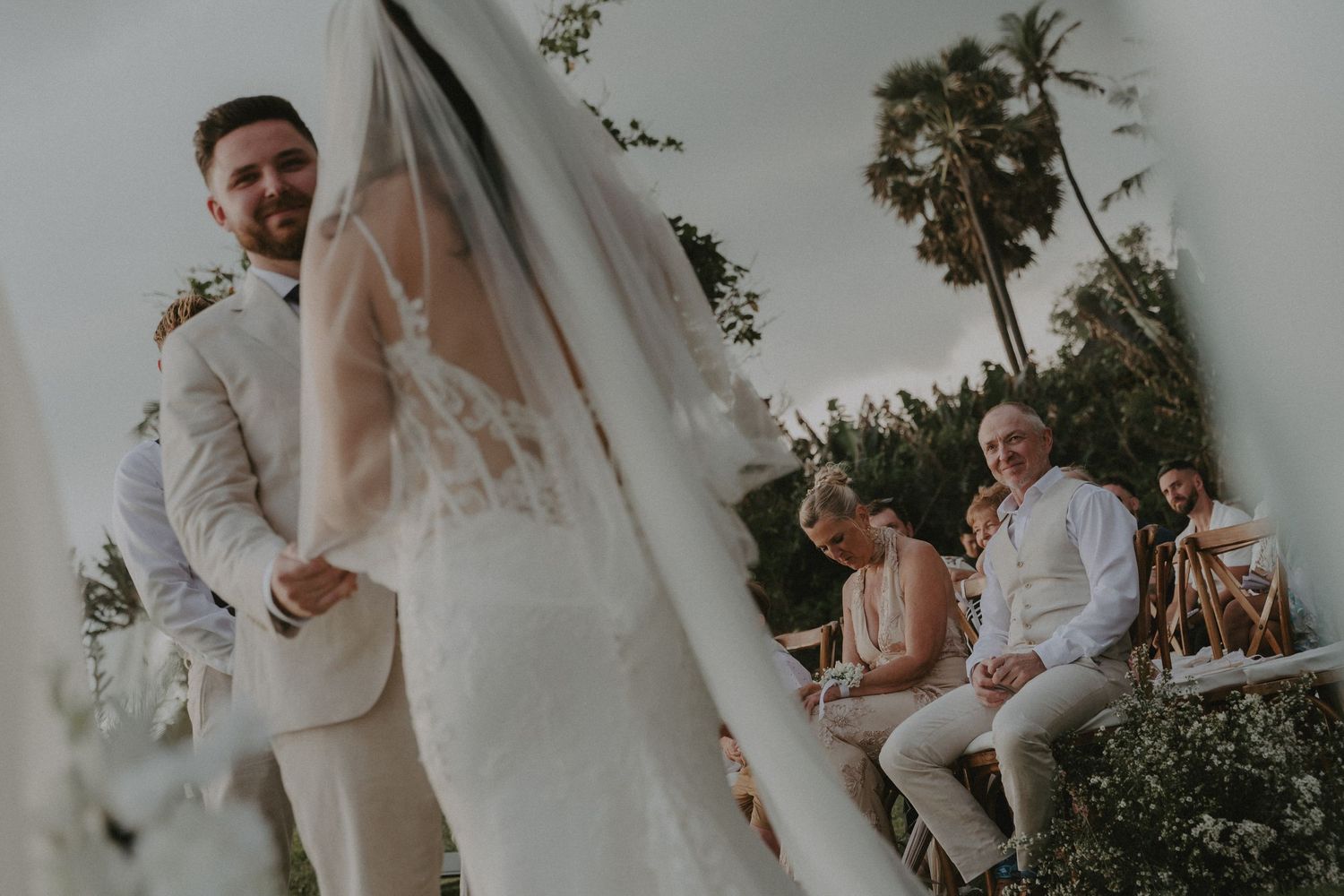 A romantic wedding ceremony under palm trees with flowing white fabric and floral decorations creating an elegant tropical setting.