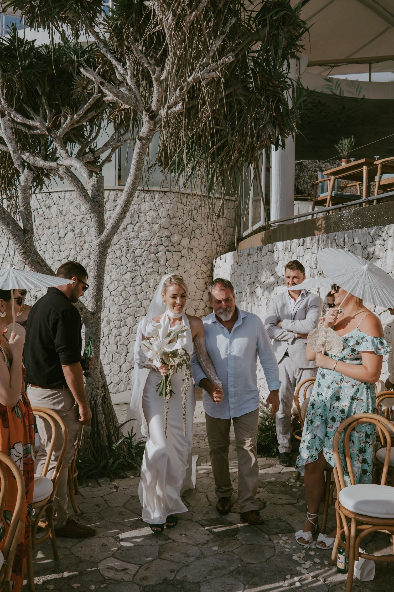 Wedding guests mingle under a covered outdoor area with stone walls and tropical foliage.