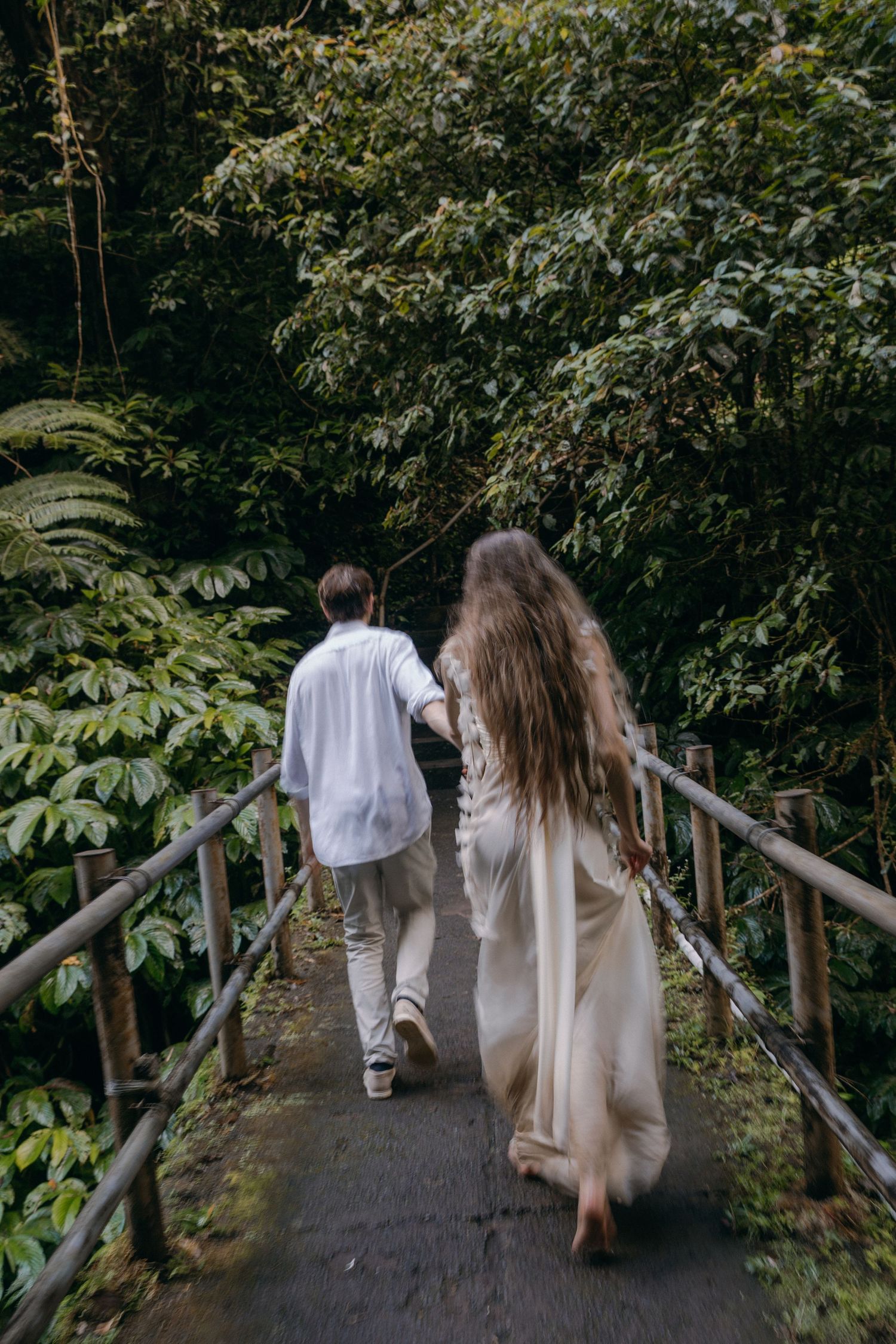 Two people stroll down a shaded garden path with wooden railings amidst dense foliage and trees.