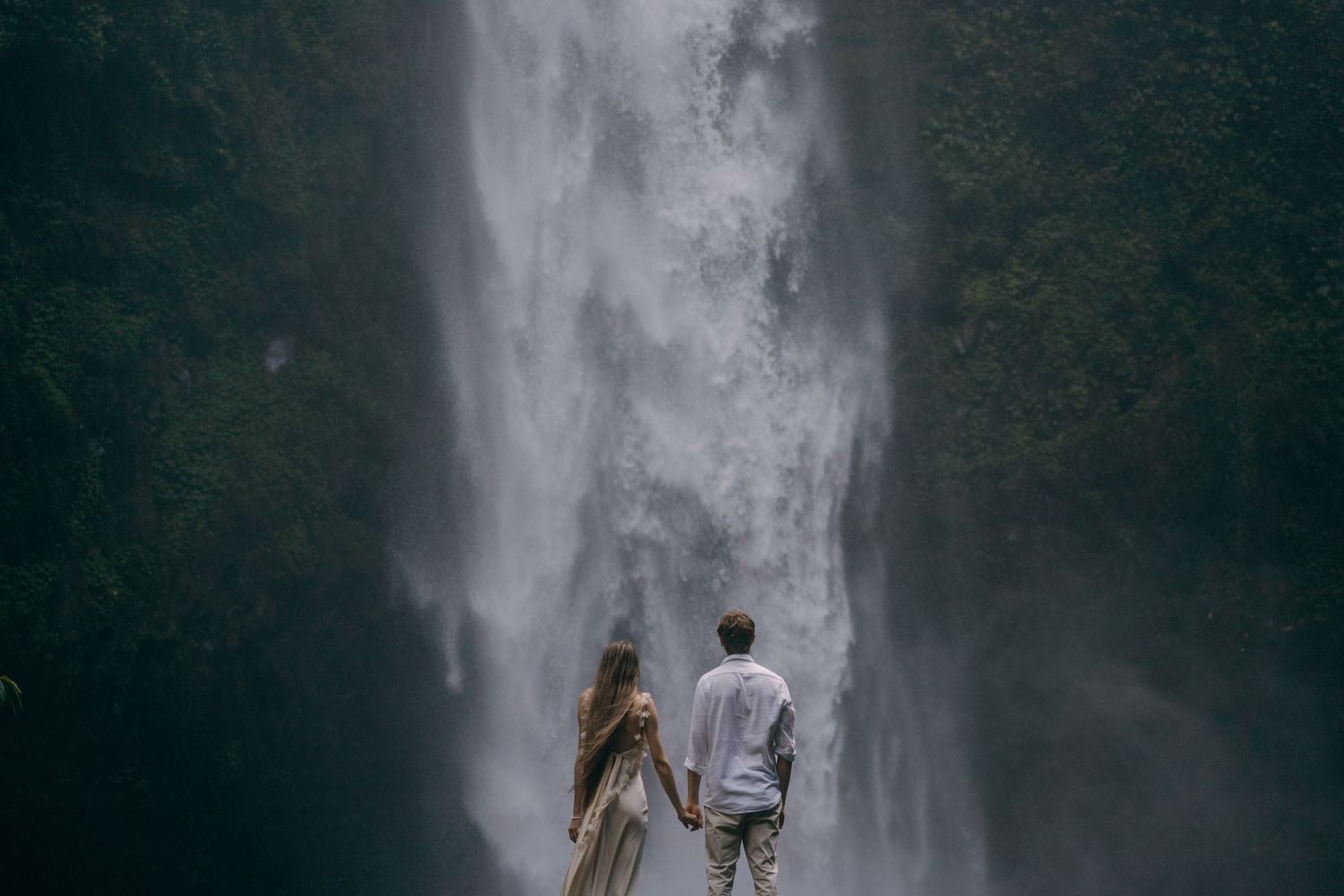 Two people stand in silhouette against a majestic waterfall backdrop in a misty forest setting.
