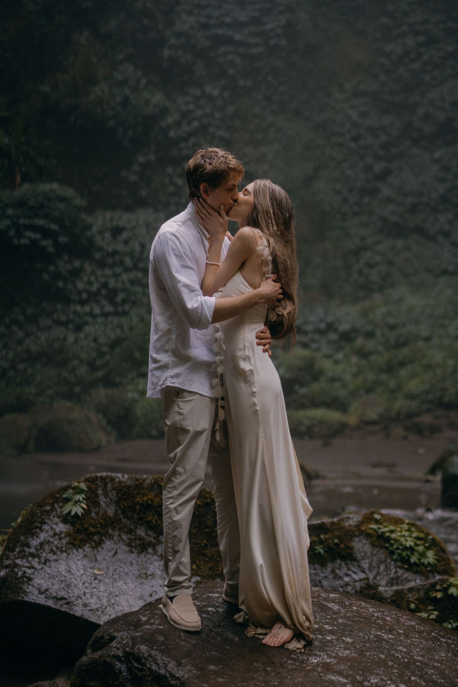 A romantic couple in white attire shares an intimate moment on a misty mountain ledge during their wedding photoshoot.