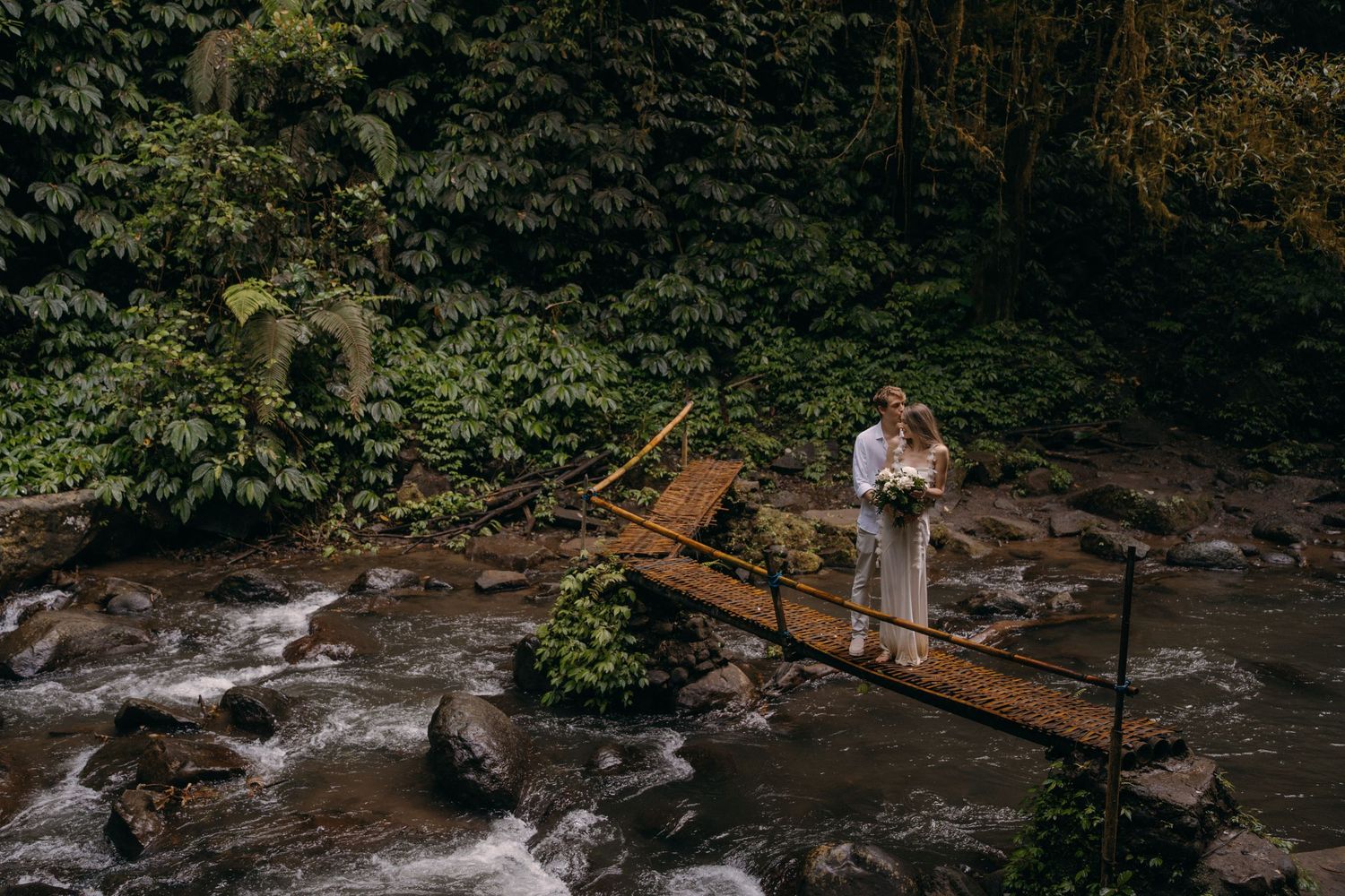 A person in white stands on a rustic wooden bridge over rushing water in a lush forest setting.