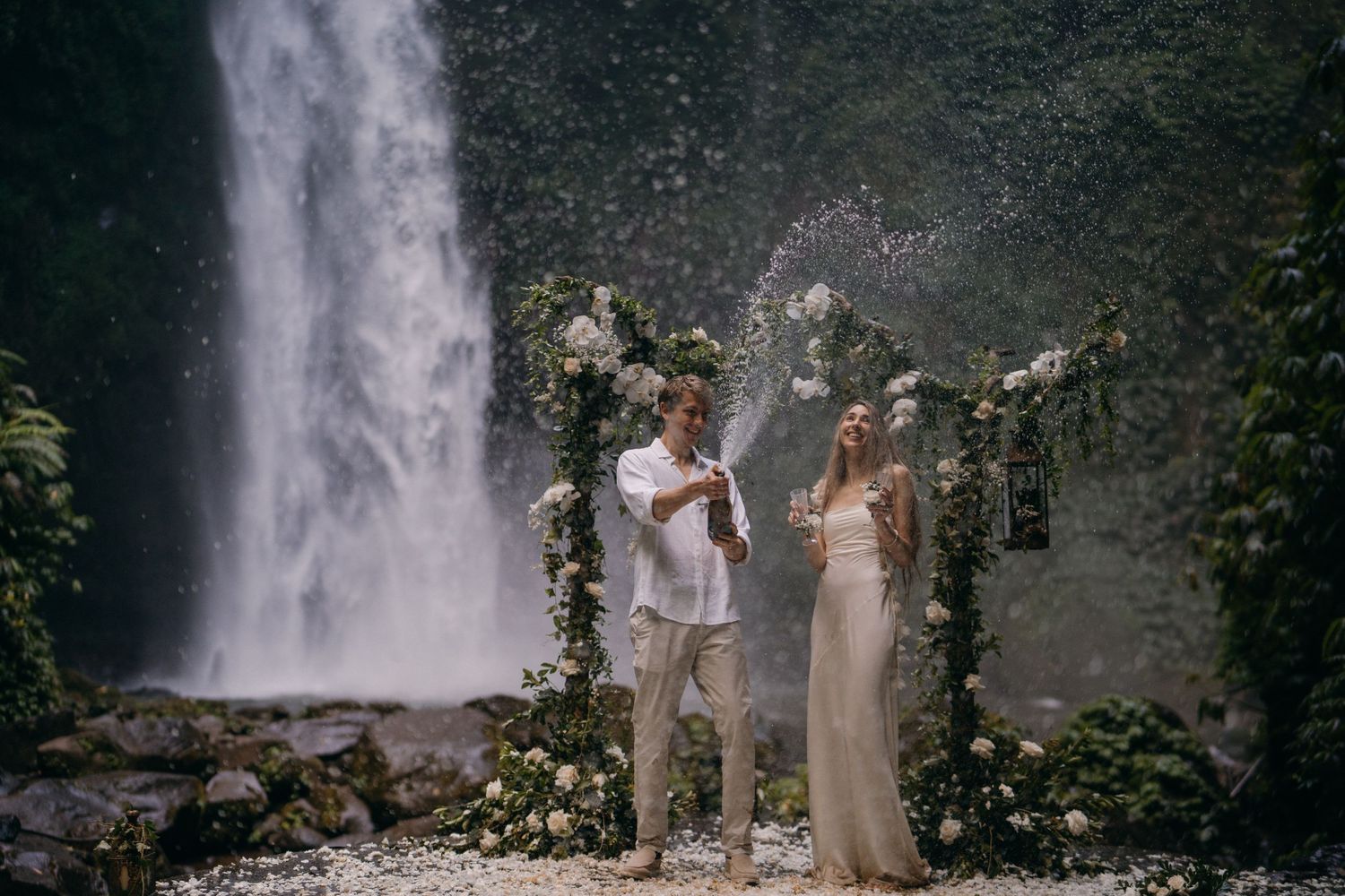 A group gathers near a scenic waterfall in a lush forest setting for what appears to be a wedding ceremony.