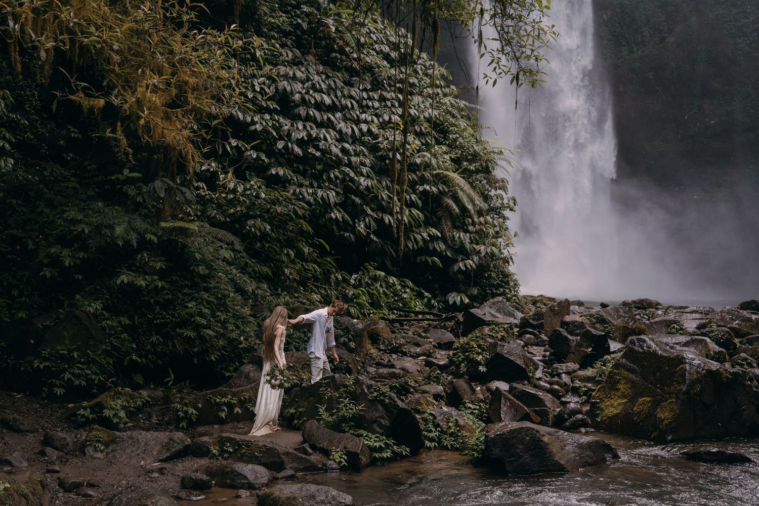 Misty waterfall cascades down moss-covered rocky cliffs in a lush tropical rainforest setting.