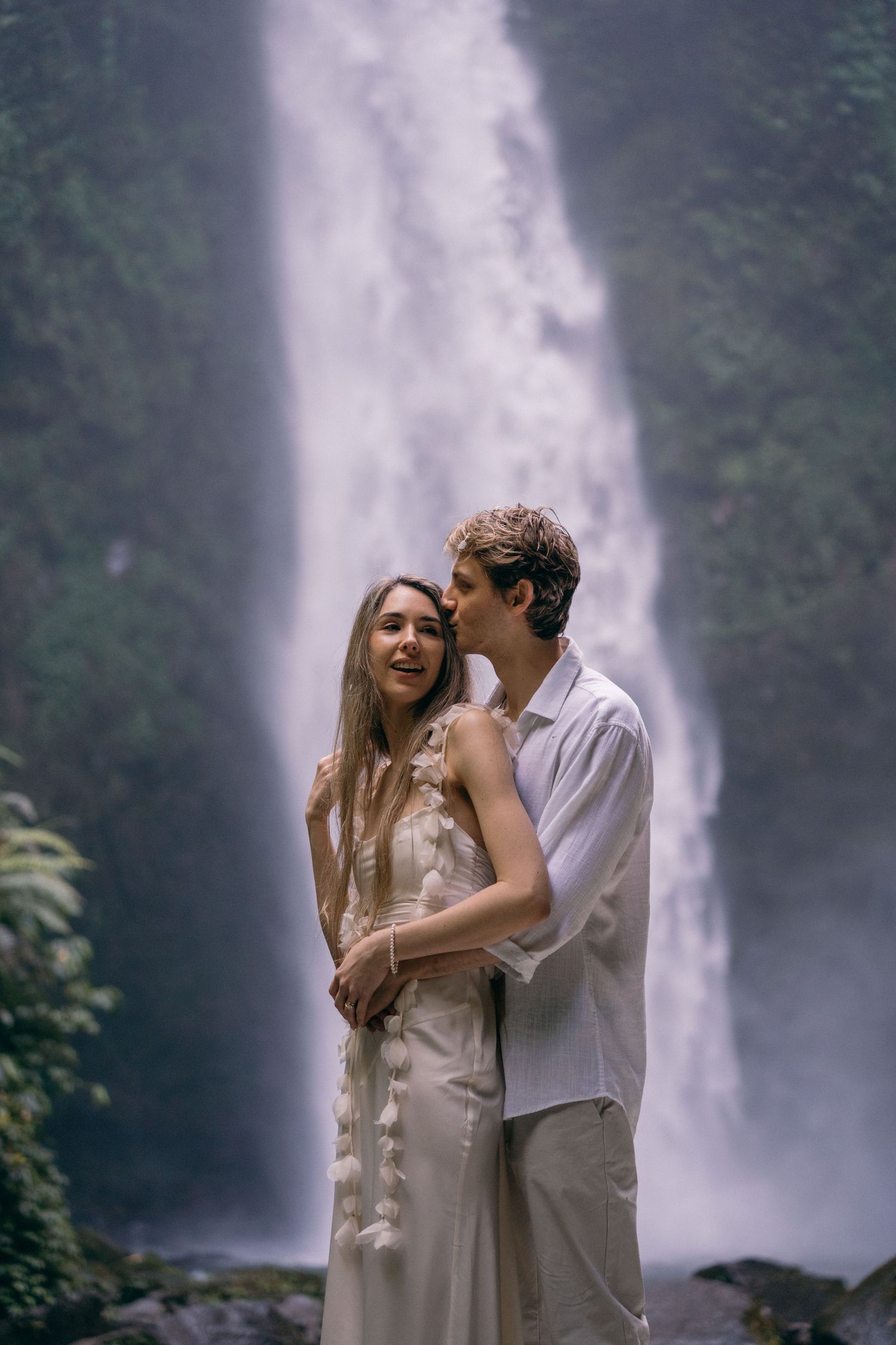 A romantic couple embraces in front of a majestic waterfall surrounded by lush greenery in an ethereal nature setting.