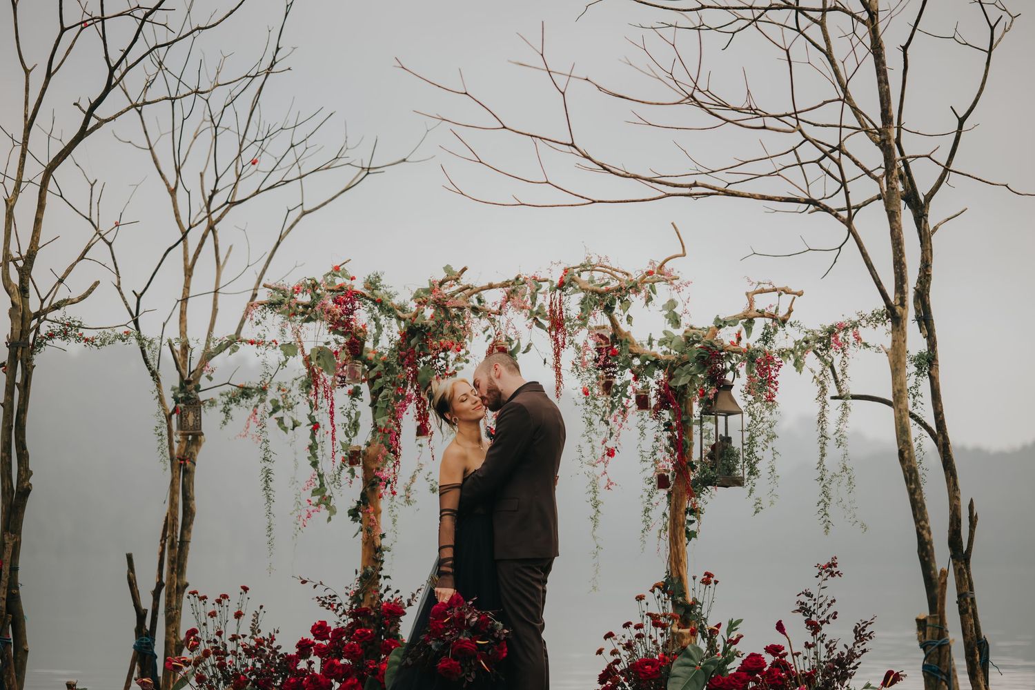 A romantic couple stands among bare winter trees with red flowering vines in the background.