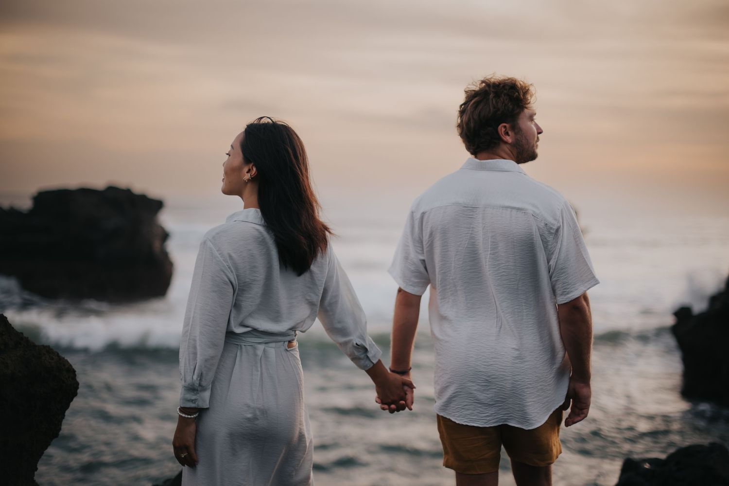 A couple holds hands while walking along a rocky beach at sunset with dramatic coastal scenery in the background.
